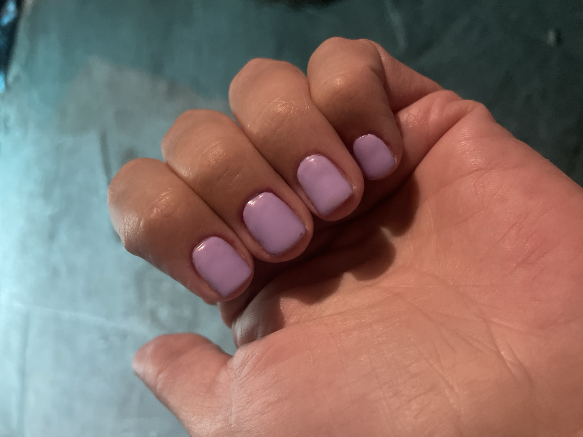 A close-up photograph shows a human hand with nails painted a light purple color. The background is blurred and out of focus, showing a light blue or gray surface with indistinct patterns. The scene does not contain enough information to place it in Olginka, Russia, or any other specific location.