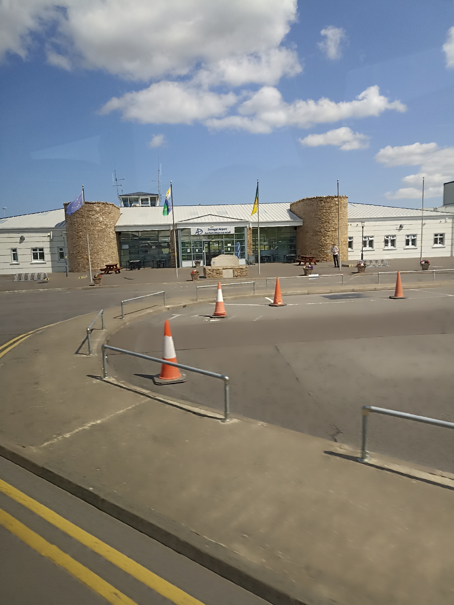 This is a daytime, outdoor, medium-distance, eye-level shot of the Donegal Airport terminal building, located in Carrickfinn, Ireland. The building is modern with a white, sloped roof and a facade featuring large glass windows and two circular stone towers. An EU flag and a green and yellow flag are flying from poles in front of the building. A man is standing near the entrance. In the foreground, there is an asphalt road with yellow double lines, and a sidewalk with metal railings. Several orange traffic cones are placed on the road and sidewalk, suggesting ongoing work or traffic management. The sky is blue with scattered white clouds.