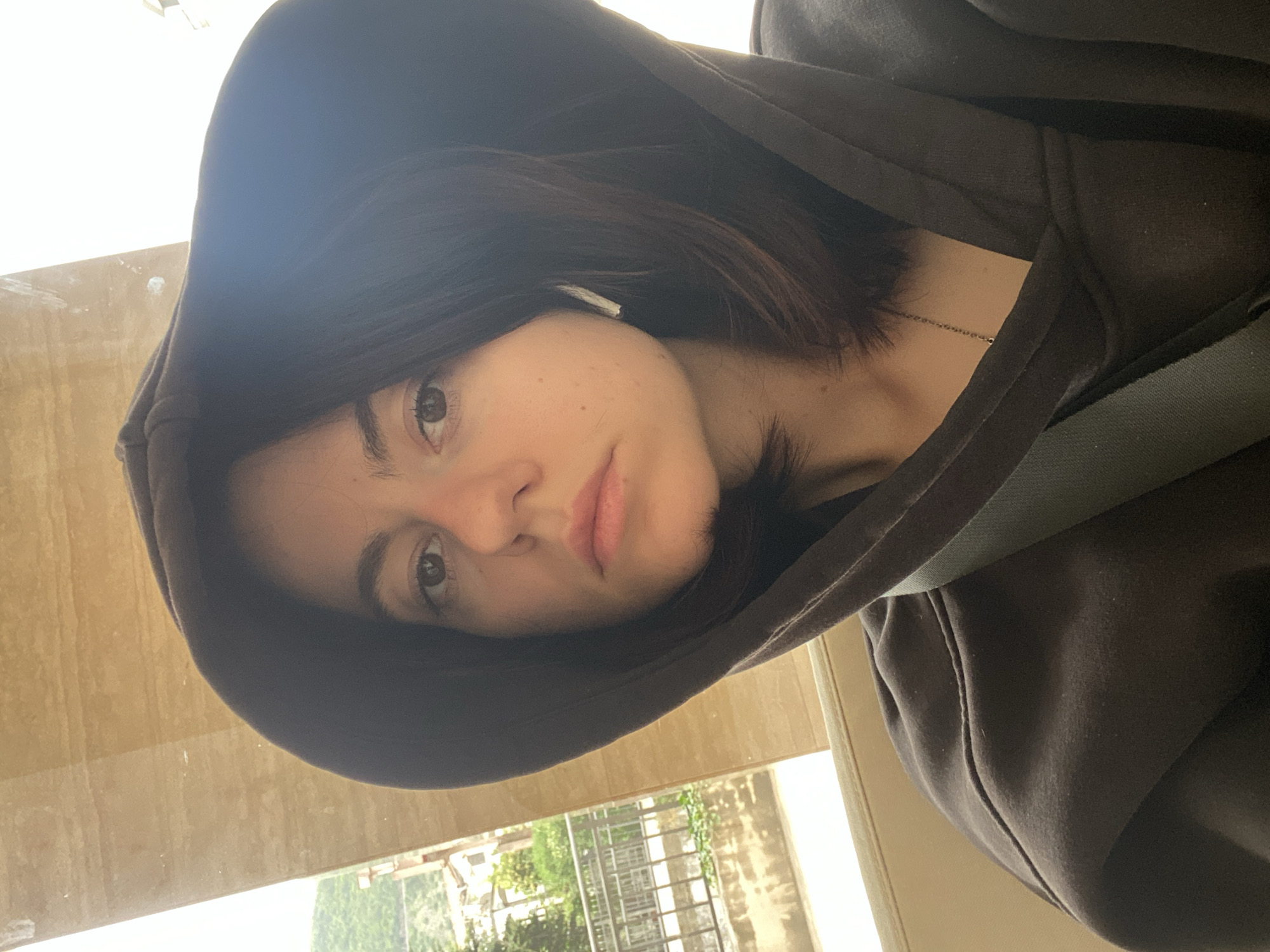 A woman with short brown hair is wearing a dark hoodie with the hood up, revealing a white earbud in her left ear. She appears to be in a vehicle, wearing a seatbelt. Outside, a view through the window reveals trees, buildings, and what appears to be a metal staircase or balcony. The location is given as "Unknown, Unknown".