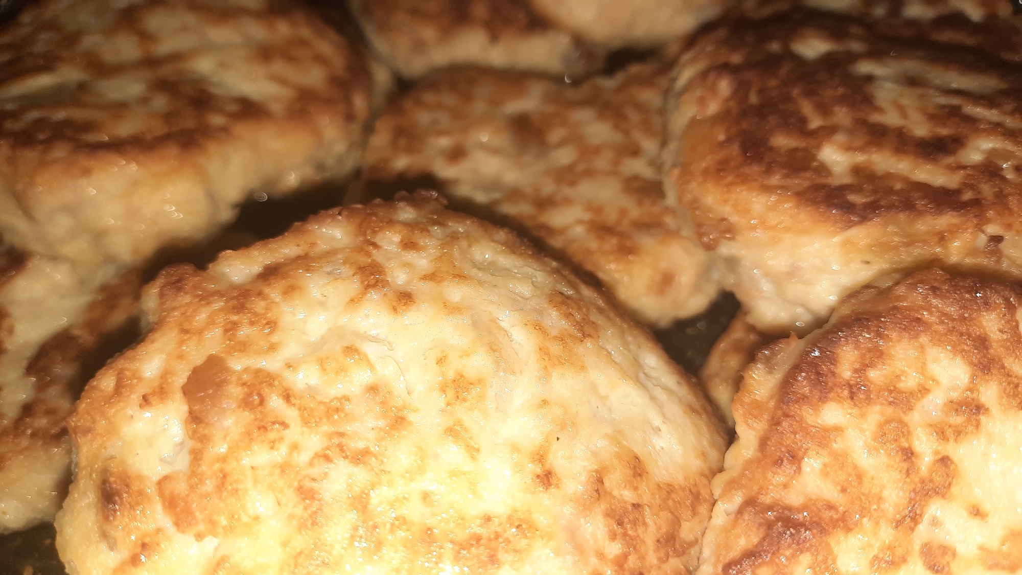A close-up, overhead shot displays a pile of golden-brown chicken cutlets. The cutlets are round and flat, with a slightly irregular, textured surface, indicating they have been pan-fried. Some areas show a deeper, caramelized brown, while others are a lighter golden hue. The lighting is warm, casting subtle shadows and highlights that emphasize the texture of the food. The background is dark and out of focus, making the cutlets the sole subject of attention. There is no indication of people, action, or an environment beyond the food itself. The scene is devoid of any context related to the city of Шклоў, Belarus, as the image focuses exclusively on the culinary subject.