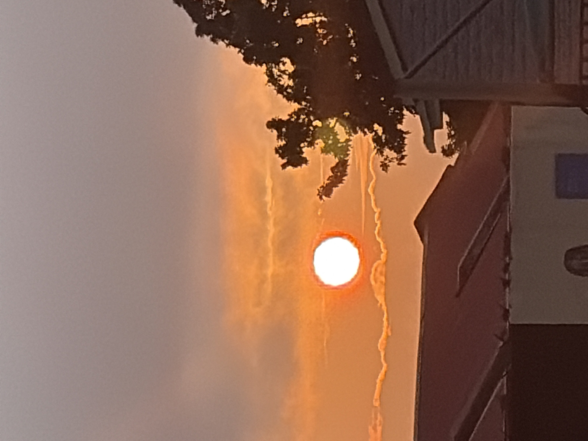 This image captures a sunset in Gura Top, Nigeria, with the sun appearing as a bright white orb with an orange halo, set against a sky of pale pink and orange hues. The sun is positioned slightly left of center, casting a warm glow on the scattered, wispy clouds below. In the foreground, the dark silhouettes of rooftops from residential buildings are visible, with one roof appearing to be made of dark red corrugated material. To the right of the frame, the dense foliage of a tree, also in silhouette, partially obscures a brick-patterned structure. The overall scene depicts a calm evening at dusk.
