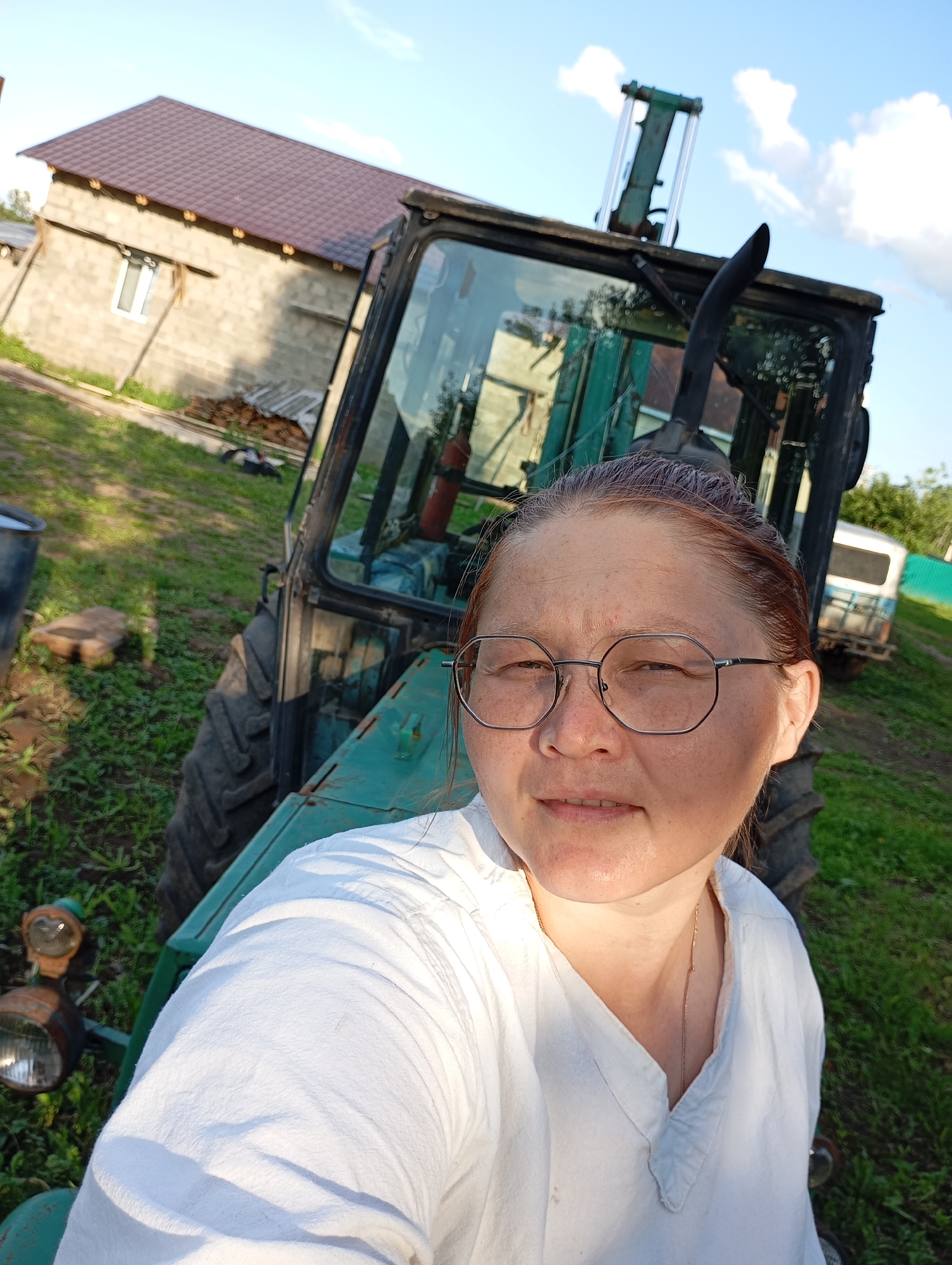 A close-up selfie taken outdoors shows a woman with glasses and her hair pulled back, wearing a white shirt. She is positioned in the foreground, with a green tractor and a building in the background. The environment appears to be rural, with green grass and a blue sky with clouds. The location is identified as Ryabki, Russia.