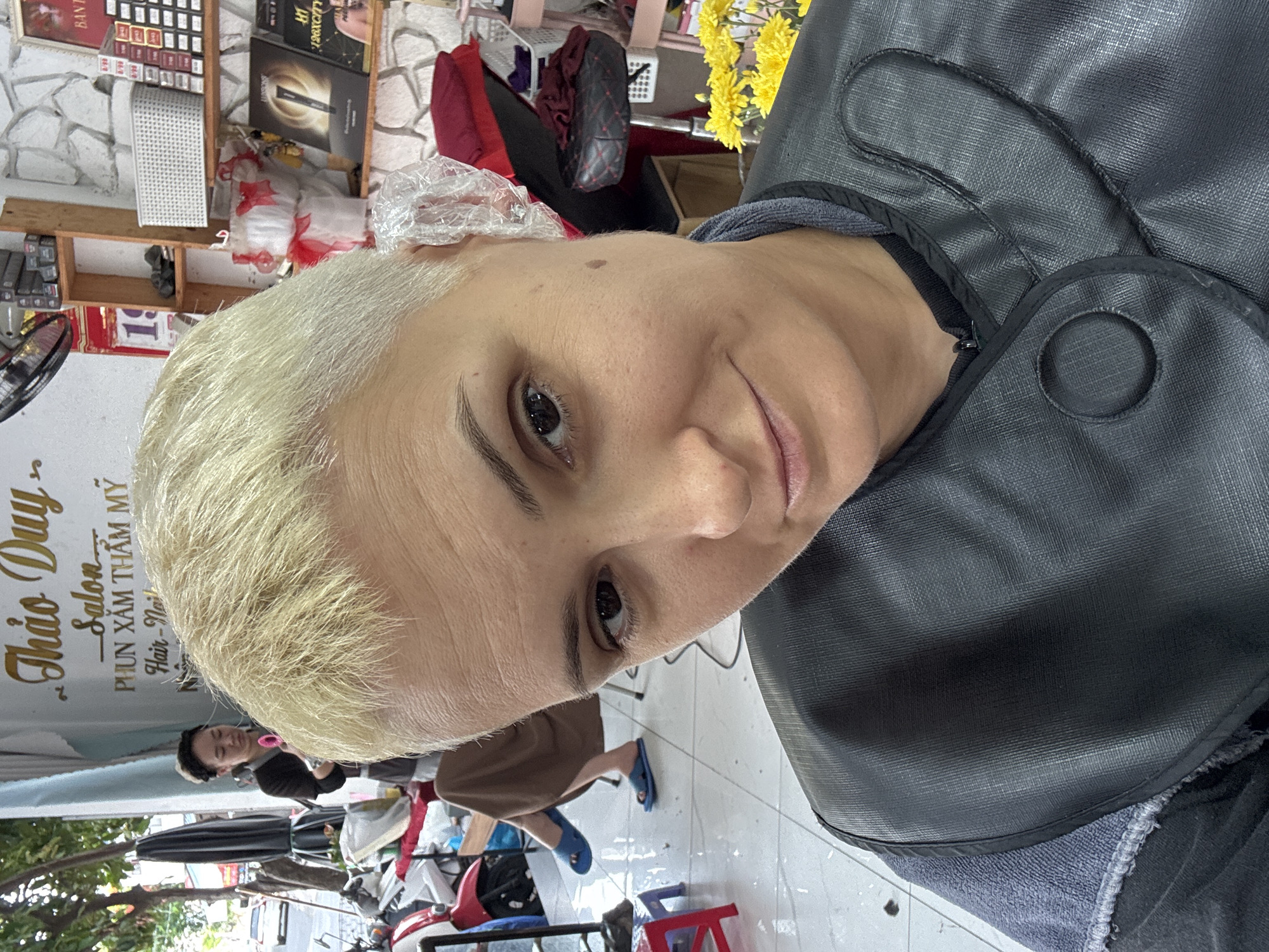 The image depicts a close-up of a person with short, bleached blonde hair, wearing a black salon cape. The individual is looking directly at the camera with a slight smile. Behind them, a salon environment is visible, with shelves containing hair products, a calendar with the number "19" on it, and decorative items. To the left, another person, presumably a salon worker, is visible wearing a black shirt and holding what appears to be a pair of scissors and a comb, with a motorcycle and a red stool in the background. The floor is tiled, and a bouquet of yellow flowers is present. The signage above the salon worker's head reads "Thao Duy Salon Phunxam Thammmy Hair - Nail", indicating the location is a beauty salon in Nha Trang Ward, Vietnam.