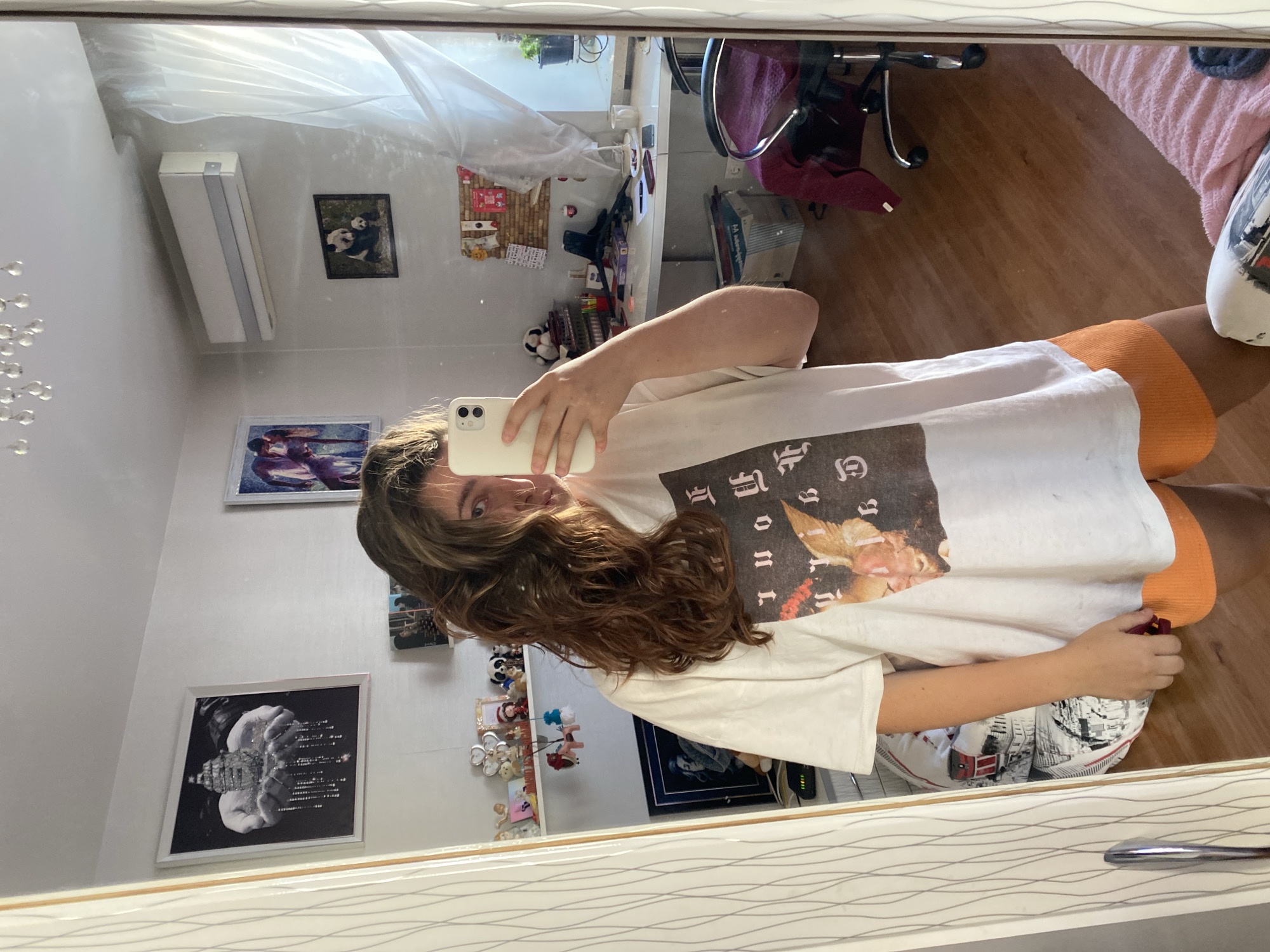 A young woman with long, brown, wavy hair takes a mirror selfie in a room. She is wearing an oversized white t-shirt with a graphic print and orange shorts. Her right hand is holding a white smartphone, and her left hand is resting by her side. The room appears to be a bedroom or living space, with a wooden floor and light-colored walls. In the background, reflected in the mirror, are various objects, including pictures on the wall, a desk with a computer and office supplies, shelves with knick-knacks, and a chair. There is also an air conditioning unit mounted on the wall and a large window with sheer curtains. The overall lighting suggests it is daytime. The location is not identifiable as Perm, Russia.