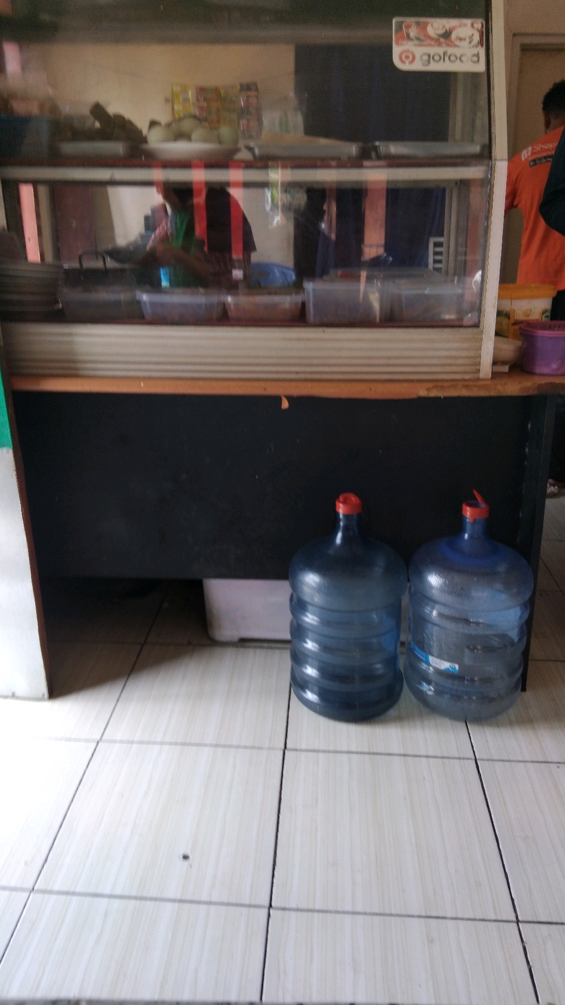 In Batang, Indonesia, a glass-fronted display case sits atop a black counter. Inside the case, various food items and containers are visible, including what appears to be boiled eggs in a bowl, and stacks of food containers. A logo for "gofood" is affixed to the upper right corner of the display case. To the right of the counter, a person wearing an orange shirt stands, partially visible. On the floor in front of the counter, two large blue water jugs with red caps are placed side by side. The floor is tiled with a light-colored, wood-grain pattern. A white plastic container is situated behind the water jugs.