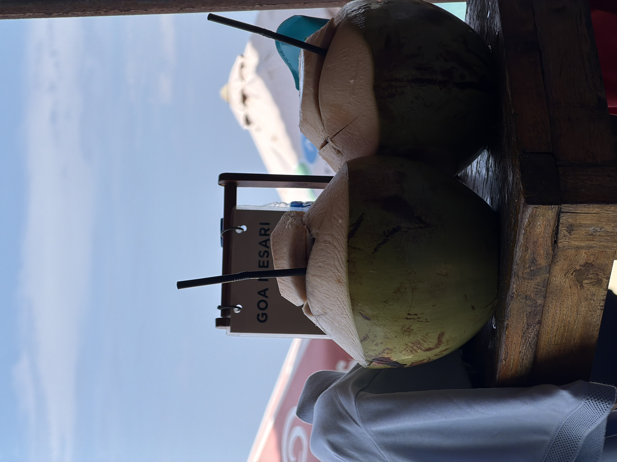 Two whole green coconuts, each with a black straw inserted, sit on a rustic wooden table. The coconuts are prepared for drinking, with the tops cut open and a piece of coconut flesh placed on top of one. In the background, a sign reads "GOA MESARI". Beyond the sign, a bright blue sky with scattered clouds is visible, along with the tops of white beach umbrellas. A light blue garment is draped over the left side of the table, partially obscuring the leftmost coconut. The scene appears to be outdoors, likely at a beachside establishment in Kuta Selatan, Indonesia, given the presence of beach umbrellas and the sign.