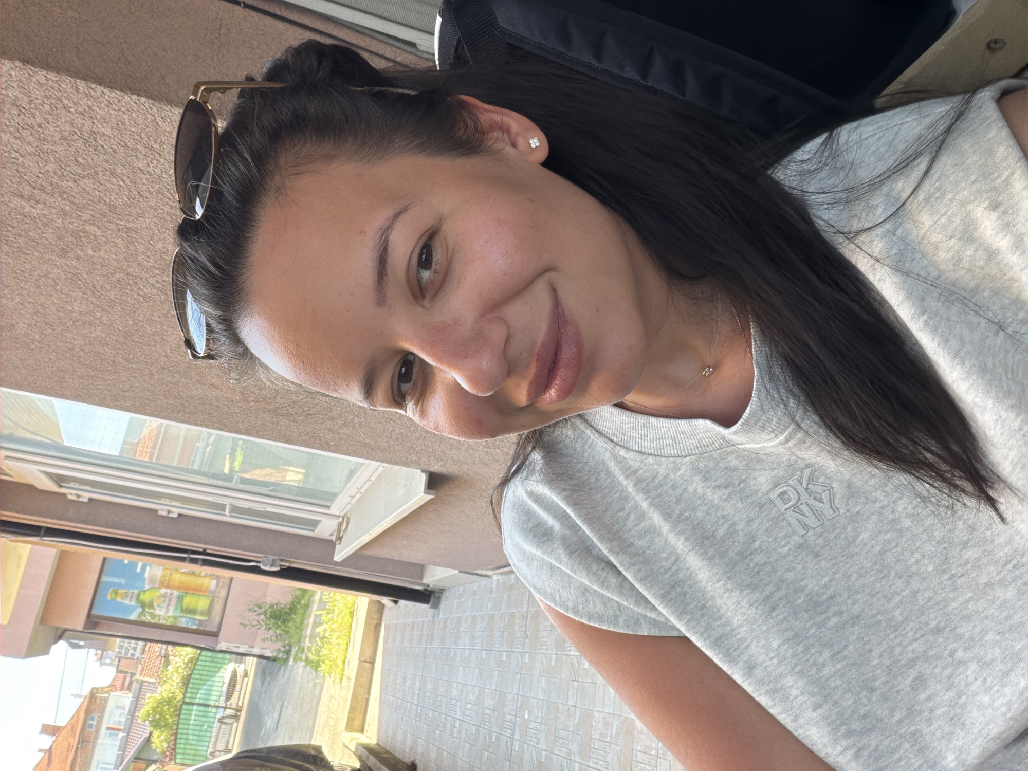A woman with long, dark hair is captured in a close-up, slightly tilted selfie. She is wearing a light grey t-shirt with a subtle "DKNY" logo embossed on the left chest. A delicate silver necklace with a small pendant is visible around her neck. She has a pair of gold-rimmed sunglasses perched on top of her head. Her expression is a gentle smile, and she appears relaxed.
The background suggests an outdoor setting. To the left, a light brown textured wall is visible, possibly part of a building. Below this, a window with white frames is open, revealing a reflection of a billboard or advertisement with two bottles of beverages. Further into the background, there are glimpses of buildings with terracotta roofs and green foliage, indicative of a residential or urban area. The ground in the foreground is tiled with a light grey and beige pattern. The overall lighting suggests it is daytime, possibly late afternoon, with soft sunlight illuminating the scene and casting subtle shadows. The setting appears to be Burgas, Bulgaria.