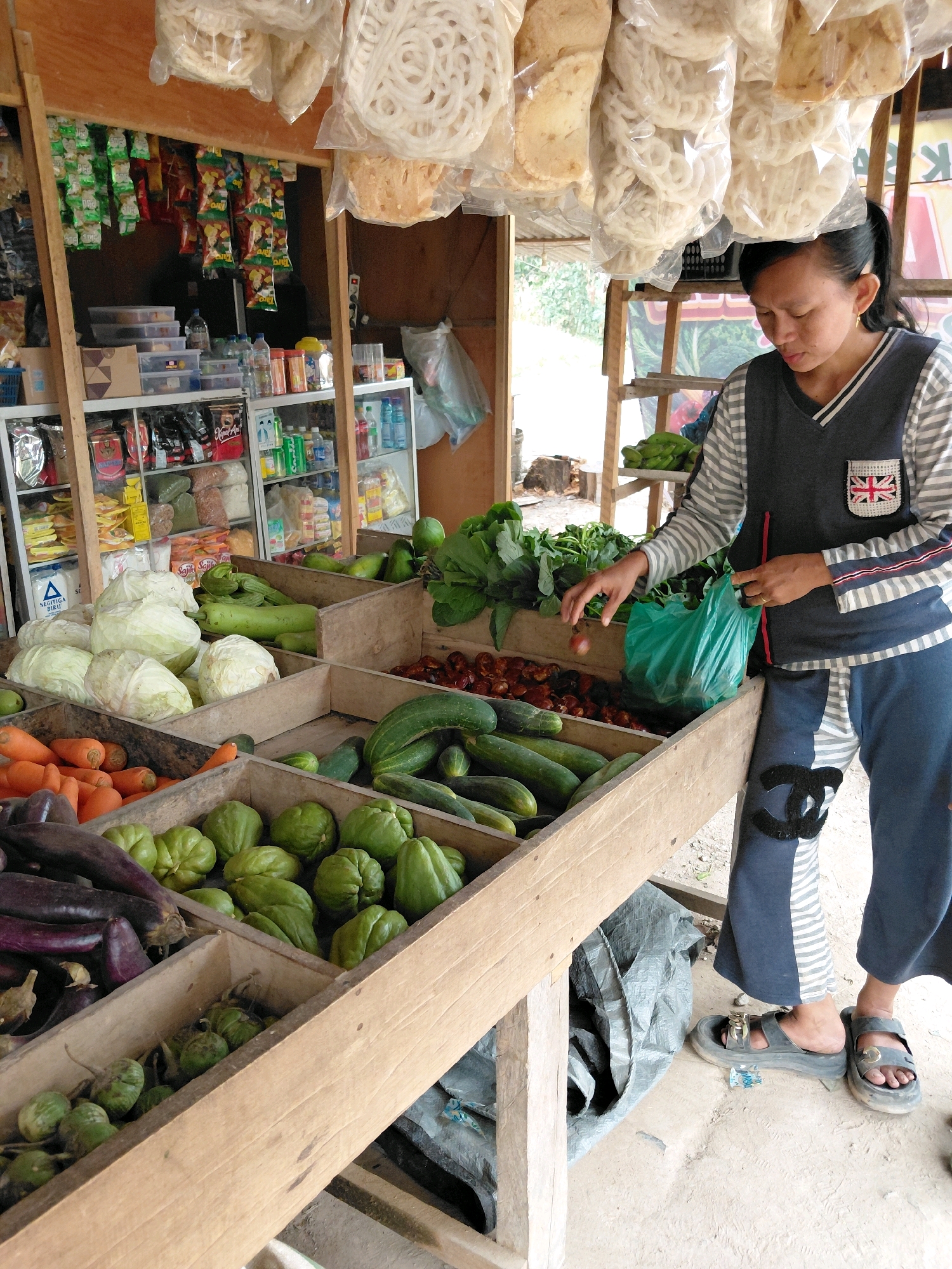 A female vendor with dark hair pulled back, wearing a grey and white striped long-sleeved shirt with a dark vest over it and dark pants with a black Chanel-like logo on the left leg, stands behind a wooden stall displaying fresh produce. The stall is filled with various vegetables, including cabbages, cucumbers, chilis, carrots, eggplants, and smaller green gourds. The vendor is holding a green plastic bag and reaching into a wooden crate of small red chilis, picking one up with her right hand. Behind the stall, shelves are stocked with packaged food items and beverages. The ground is dirt and concrete. The scene appears to be an outdoor market stall in Persiapan Mulya Raya, Indonesia.