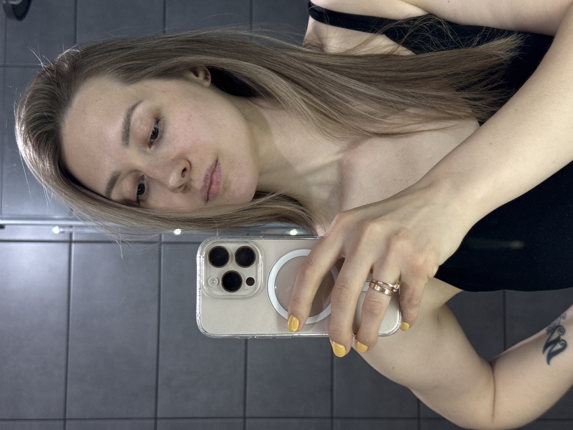 A young woman is taking a selfie in a bathroom with gray tiled walls. She's holding an iPhone in a clear case with visible camera lenses and a MagSafe ring. She has light brown hair and is wearing a black top. She has two gold rings on her finger and her fingernails are painted a yellow color. A small black tattoo is visible on her arm. Location information is not evident in the image.
