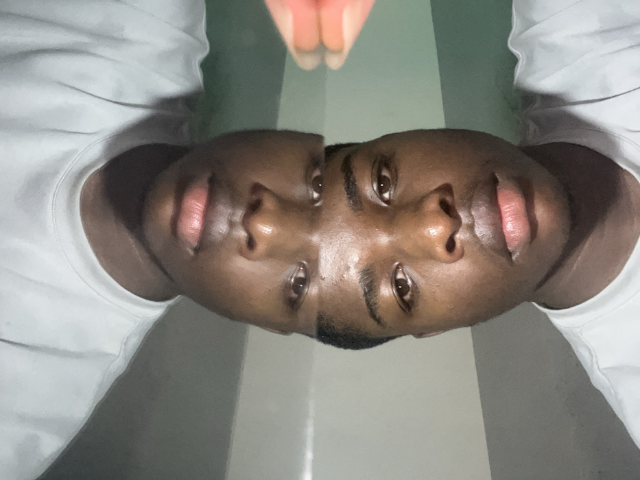 The scene depicts a young adult male located in the city of Shen, Nigeria. The individual has dark skin, short dark hair, and brown eyes, and is wearing a white crew-neck t-shirt. The image features a prominent split composition: the lower section displays his face in an upright orientation, while the upper section shows an inverted, partial reflection of his face. Both facial representations are directed forward, engaging the viewer. A reddish-orange object, consistent with a finger or part of a device, is partially visible in the upper right quadrant. The background consists of a plain wall, light grey on the left and transitioning to a greenish-grey hue on the right. No other individuals or actions are discernible within the frame.