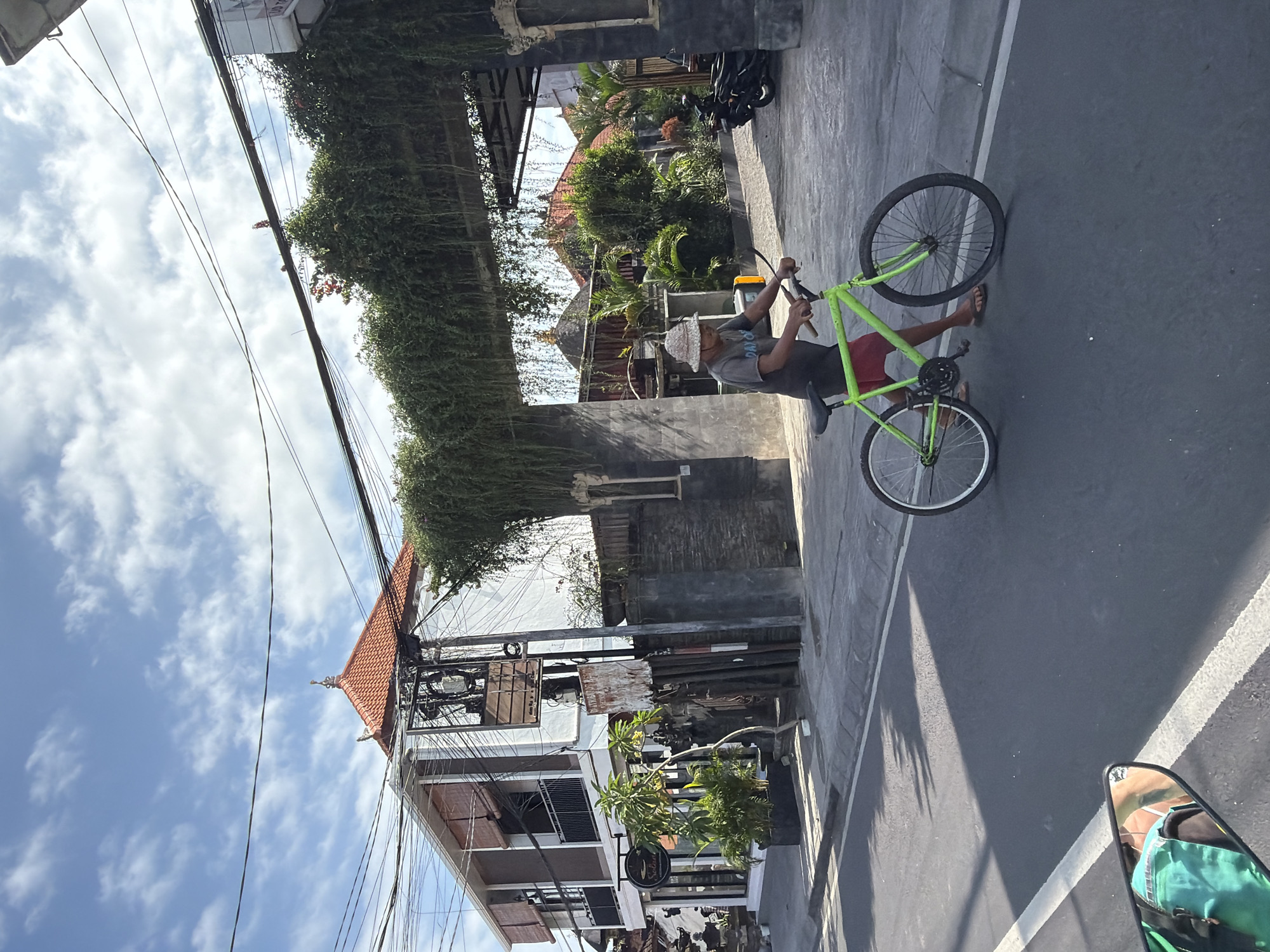 A man walks a green bicycle on a street in North Kuta, Indonesia. The man is wearing a grey t-shirt, a light-colored patterned hat, and brown sandals. The bicycle has a red rear rack. The street is paved and has a white lane marker. Buildings line the street, some with overhanging greenery. Power lines are visible against a cloudy blue sky. A motorcycle mirror reflects a masked individual on the lower right.