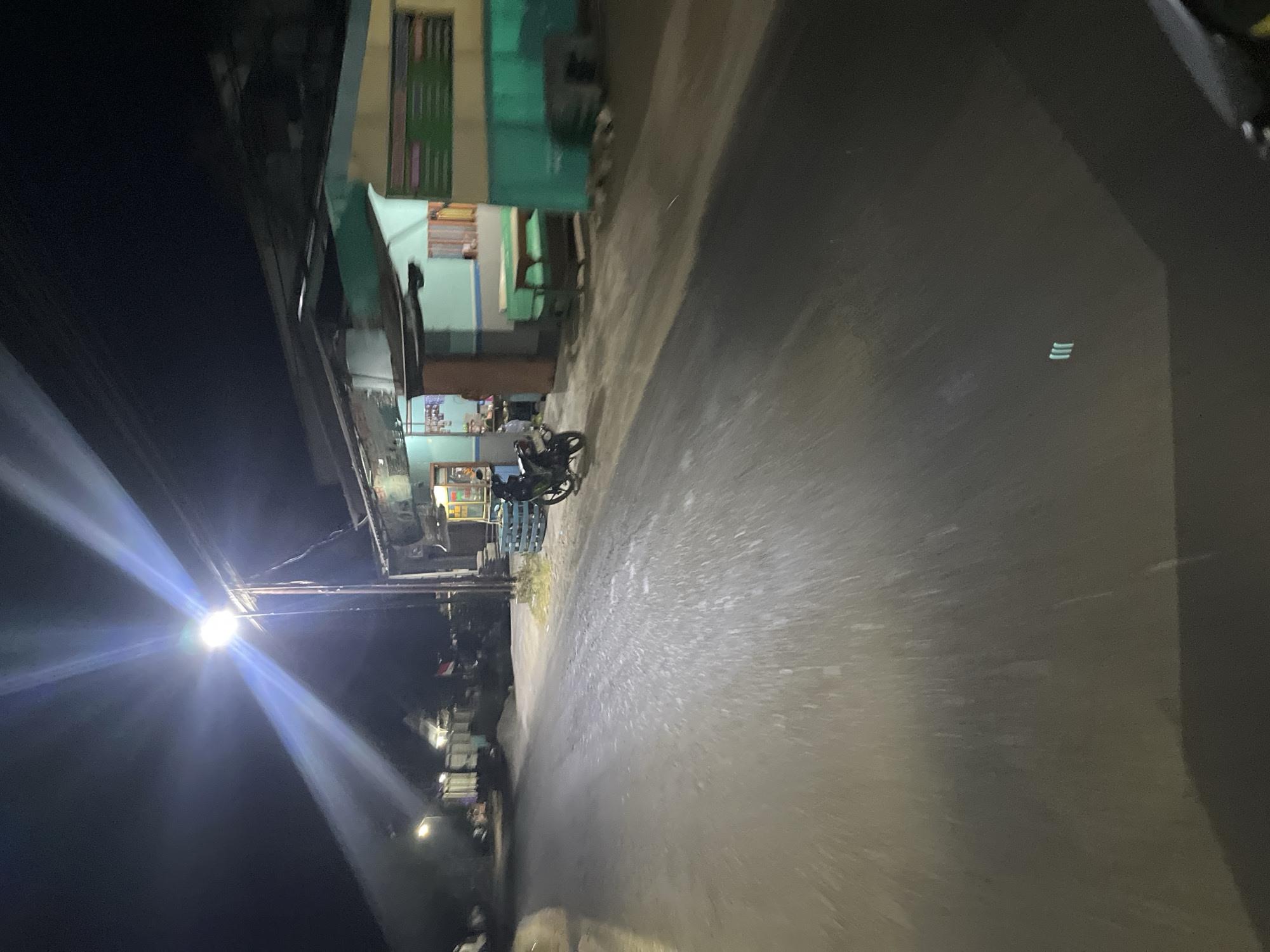 The image shows a street scene at night in Karangrandu, Indonesia. A bright street lamp illuminates the scene, casting long shadows. A motorcycle is parked on the side of the road in front of a building with a teal and yellow facade. The building appears to be a small shop or restaurant, with tables and chairs visible outside. The road is paved and appears wet, reflecting the light from the street lamp. The overall atmosphere is quiet and dimly lit, typical of a street at night in a rural or suburban area.