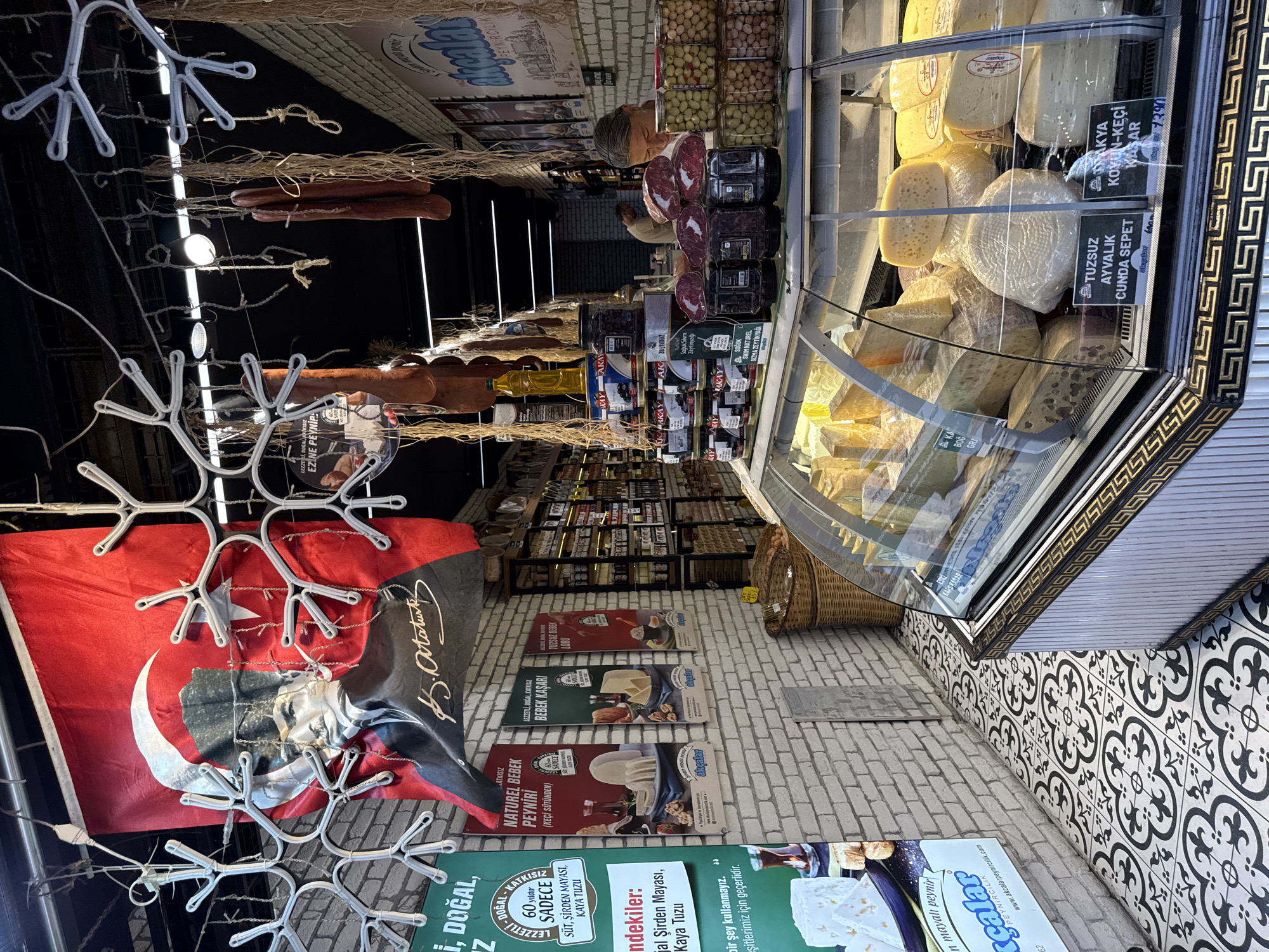 The image depicts a cheese shop in Kadıköy, Turkey, showcasing a variety of cheeses and other food products. A prominent Turkish flag with a portrait of Mustafa Kemal Atatürk hangs at the top, adding a patriotic element to the scene. Several decorative snowflake-like structures are suspended from the ceiling, along with dried meats and bunches of straw.
In the foreground, a refrigerated display case is filled with different types of cheese, some wrapped in plastic and others exposed. Labels are visible, indicating varieties like "Tuzsuz Ayvalik Cunda Sepet" and "Akyaka Koyun-Keci Kasar." Behind the display case, shelves are stocked with jars of olives, pickles, and other preserved foods. A woman with gray hair is partially visible behind the counter, interacting with the products.
The shop's interior features white brick walls and a black ceiling with visible lighting fixtures. The floor is adorned with a decorative black and white tile pattern. Posters advertising different cheese types, such as "Naturel Bebek Peyniri (Keçi Sütünden)" and "Lezzetli, Doğal, Kayıksız Bebek Kaşar," are displayed on the walls. The overall impression is that of a traditional Turkish market stall, emphasizing local produce and a sense of community.