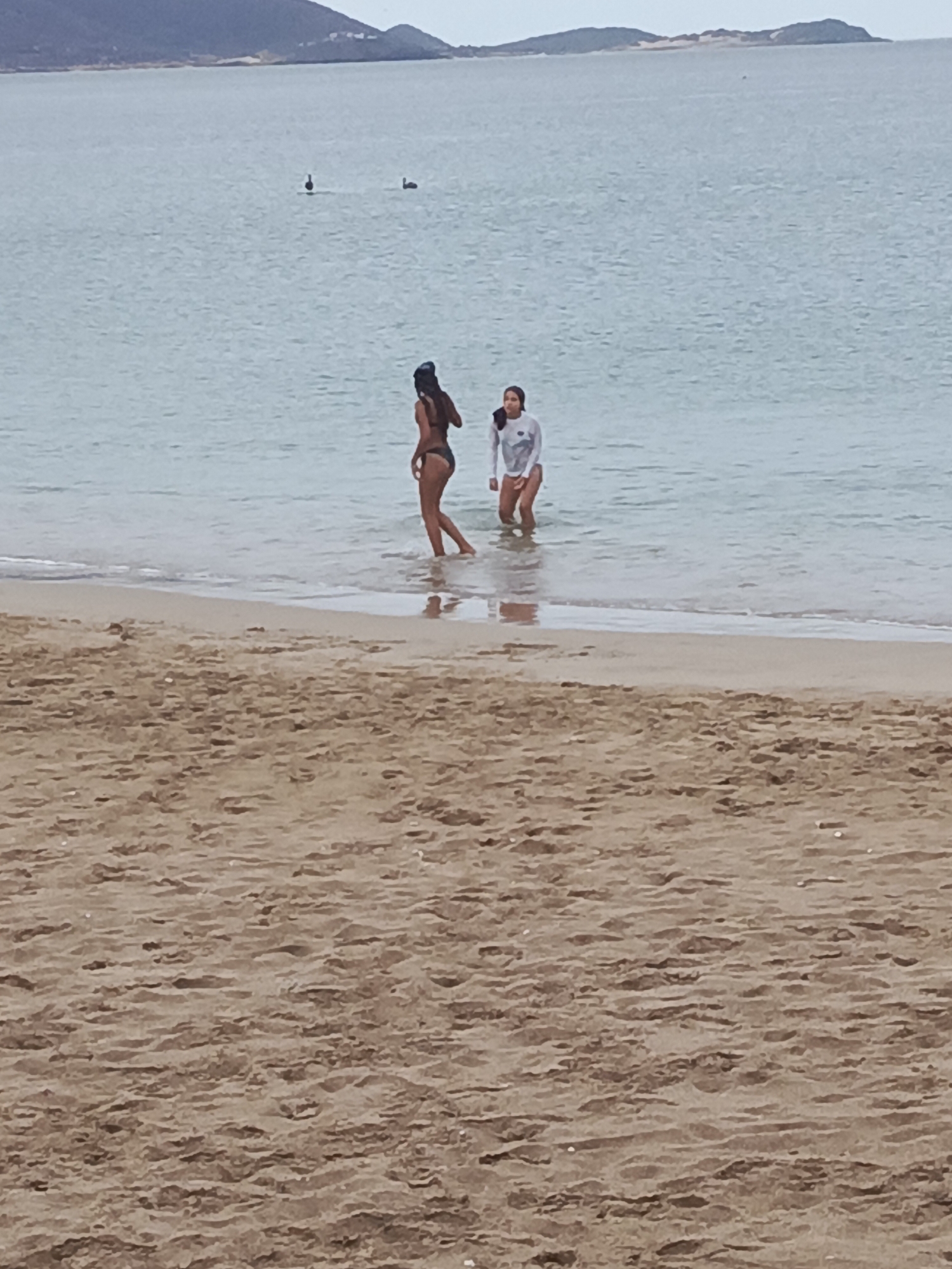 The image shows two people wading in the shallow water at a beach. The person on the left is wearing a dark bikini and has their back to the camera. The person on the right is wearing a white long-sleeved rash guard and dark bottoms, facing towards the left. They are standing in calm, light blue water. The beach in the foreground is sandy and appears to be wet from the receding tide. In the background, there is a calm body of water and a distant shoreline with hills or mountains. Two birds are visible in the water in the mid-ground, one closer and darker, the other further and lighter. The sky is overcast, suggesting a cloudy day. The scene appears to be a leisurely day at the beach. There is no visible text. The location context provided is Juan Griego, Venezuela.