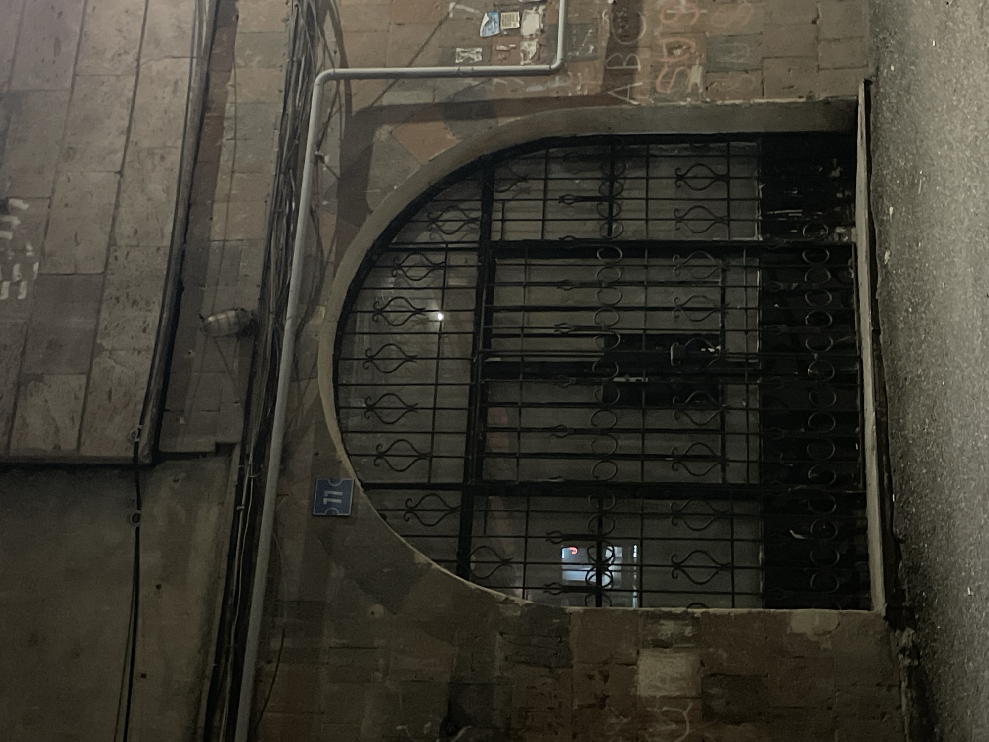A ground-level, low-angle shot shows a window with a decorative metal grate. The window is set in a brick wall with an arched top. The wall itself appears to be old, with visible wear and discoloration. To the left of the window, there are wooden panels or a door, also appearing aged and weathered. There are also conduits or pipes running along the wall. Above the window, faint inscriptions, possibly graffiti, are visible on the brick wall. A small, blue street number plaque is affixed to the wall near the window. Through the window, a glimpse of an interior space is visible, including a small illuminated object. The scene is dimly lit, with shadows indicating a nighttime setting. The location appears to be a side street or alley, and the style of architecture is consistent with older buildings in Yerevan, Armenia.