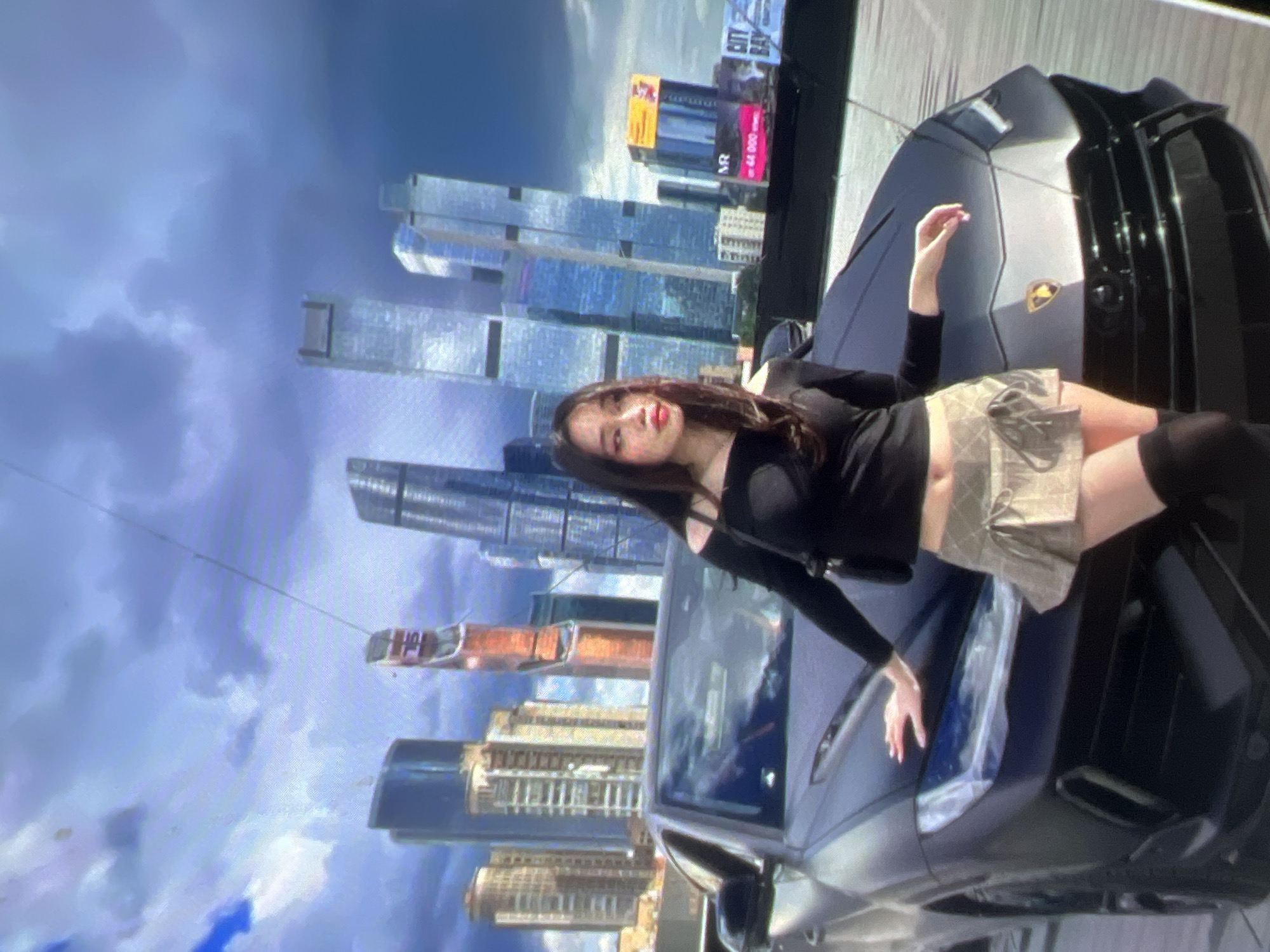 In an urban setting with a dramatic, cloudy sky, a young woman with dark hair and makeup poses beside a sleek, black Lamborghini. She wears an off-the-shoulder black top, a light-colored plaid mini-skirt, and black thigh-high socks. Her right hand rests on the hood of the car, while her left arm is extended downwards. Behind them, a cluster of modern skyscrapers pierce the sky, their glass facades reflecting the light. Billboards are visible in the background, one featuring the text "CITY BAY." The scene appears to be a posed photograph in a city, possibly an outdoor exhibition or a scenic overlook.