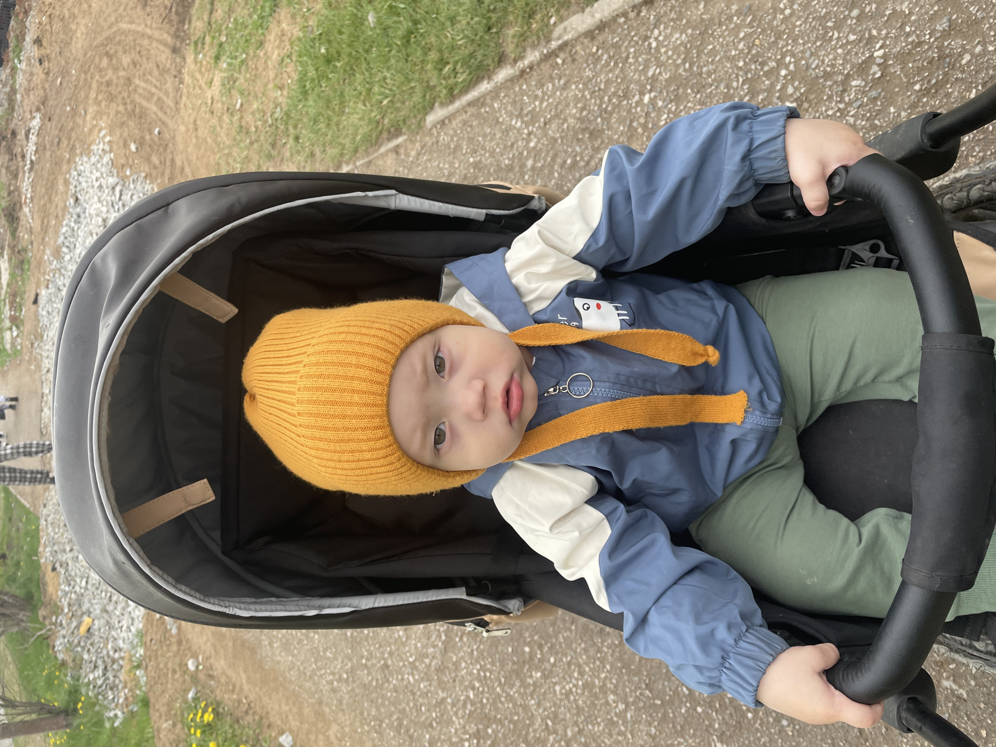A baby wearing a yellow beanie and a blue-and-white jacket sits in a black stroller. The baby is holding onto the stroller's handle. The location appears to be an outdoor setting, possibly a park or sidewalk in Perm, Russia, given the mention of the city. The ground is a mix of grass, gravel, and paved surfaces.