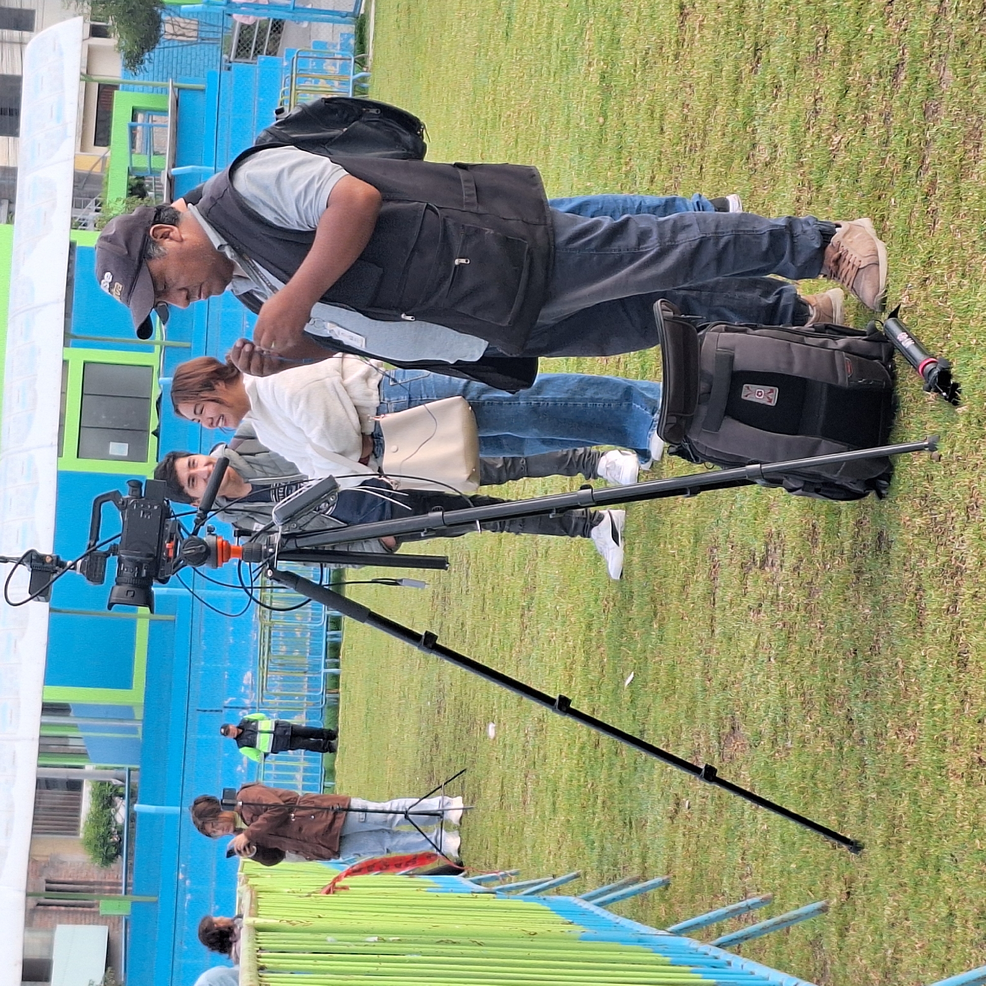 A group of people are gathered around a video camera on a tripod, apparently filming something on a grassy field. A man wearing a cap and a vest is adjusting something, while another man is behind the camera. A woman wearing a white shirt and jeans is next to them, also appearing to be involved in the filming. A camera bag is resting on the grass nearby. In the background, there are people walking, along with a building painted in blue and green. The photograph was taken in Alto Selva Alegre, Peru.