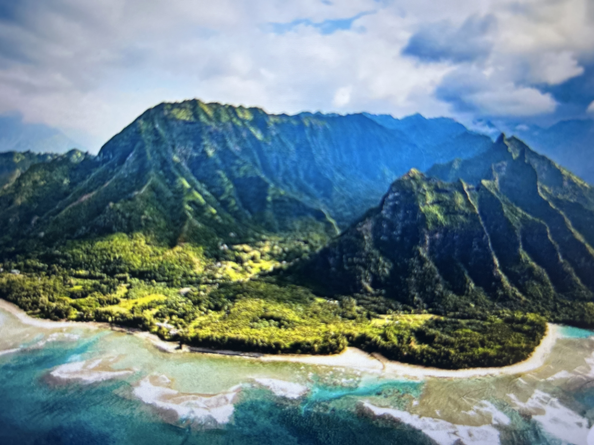 The image shows a tropical landscape with mountains covered in green vegetation meeting a coastline. The water is turquoise and tan, indicating shallow areas and possible reefs. The sky is partly cloudy. The location is not Varna, Bulgaria; the landscape resembles a tropical island like those found in Hawaii or French Polynesia.