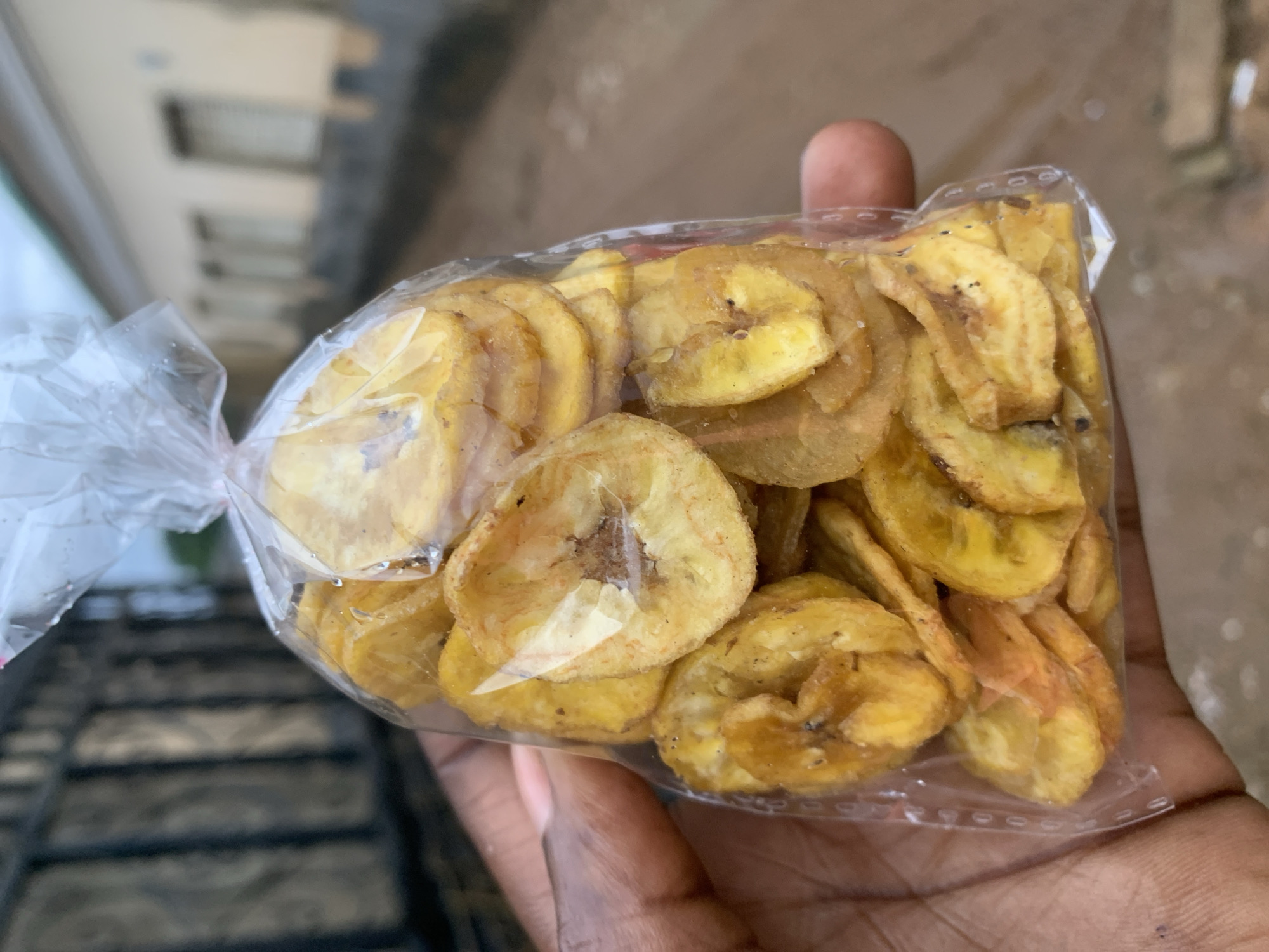 A hand holds a clear plastic bag full of banana chips. The chips are golden yellow and round. The background shows a blurred cityscape. The location is near New Stadium, Nigeria.