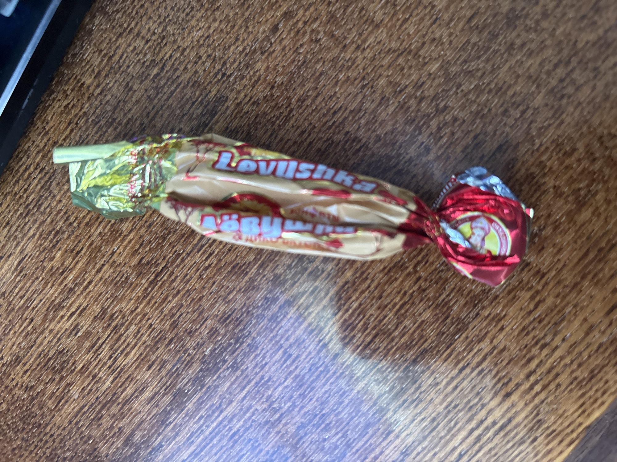 A single candy wrapped in crinkled gold and red foil rests on a wooden surface. The candy is oriented vertically, with the red foil end at the bottom and the gold foil end at the top. The gold foil is twisted at the top, revealing a small, pale green stick-like object. The wrapper has red lettering that appears to say "Lovushka" repeated. A circular logo with yellow and red is visible on the red foil at the bottom. The wooden surface has a distinct grain pattern with diagonal lines and appears to be a table or desk. The lighting suggests an indoor environment, with a shadow cast to the left of the candy. There are no people visible in the image. The location is described as being in the city of Murmansk, Russia.