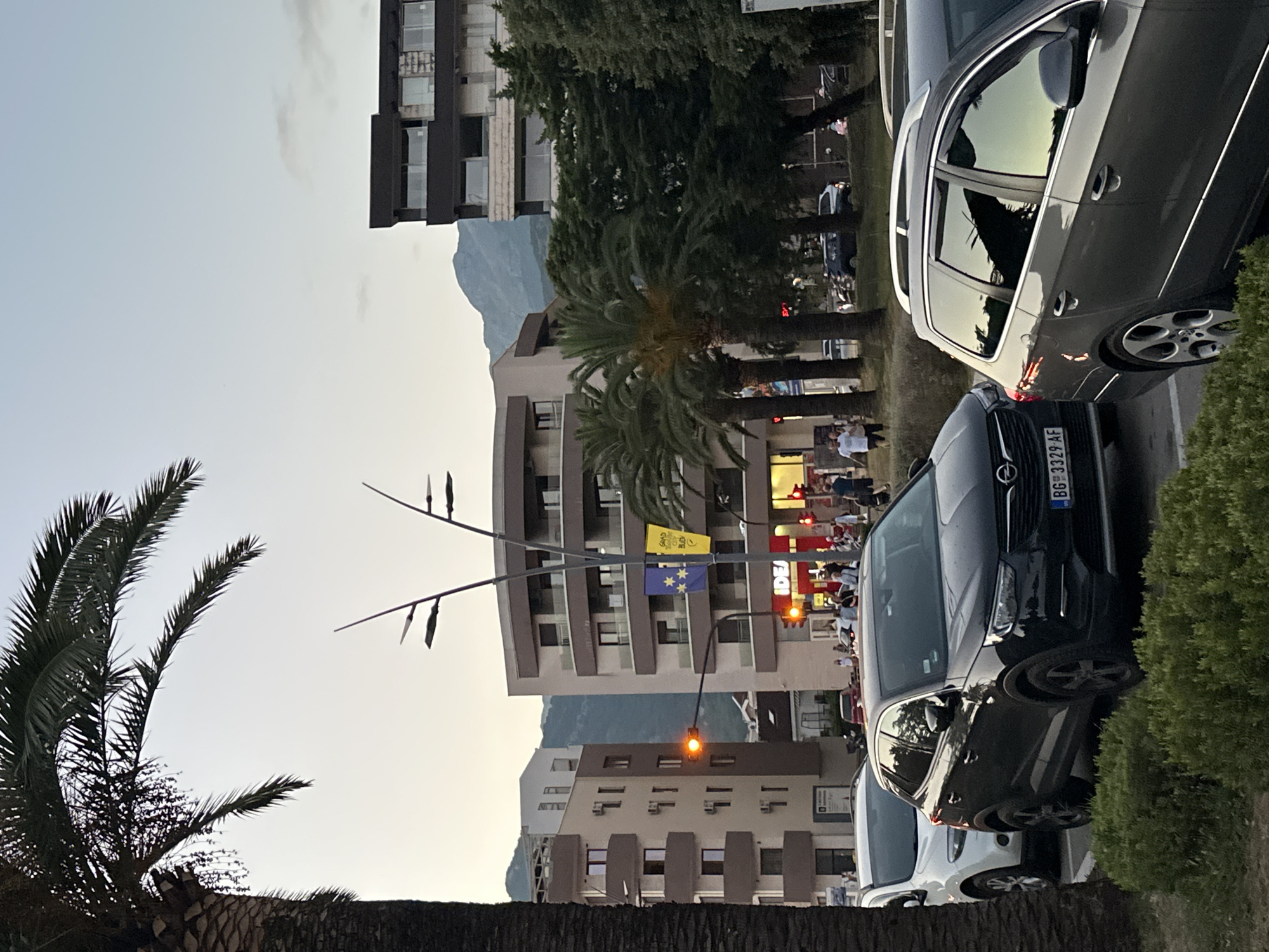 The image depicts a street scene in Budva, Montenegro, as dusk settles. A prominent palm tree is on the left, partially obscuring modern buildings. Several cars are parked along the street, including a black Opel SUV with a license plate reading "BG 3329 AF," a white car, and a gray station wagon. Pedestrians are visible walking on the sidewalk and near traffic lights. Buildings in the background include multi-story structures with balconies, and one building has a yellow flag with blue stars. A distant mountain range is visible against the twilight sky. The overall atmosphere suggests a typical urban environment during the early evening hours.