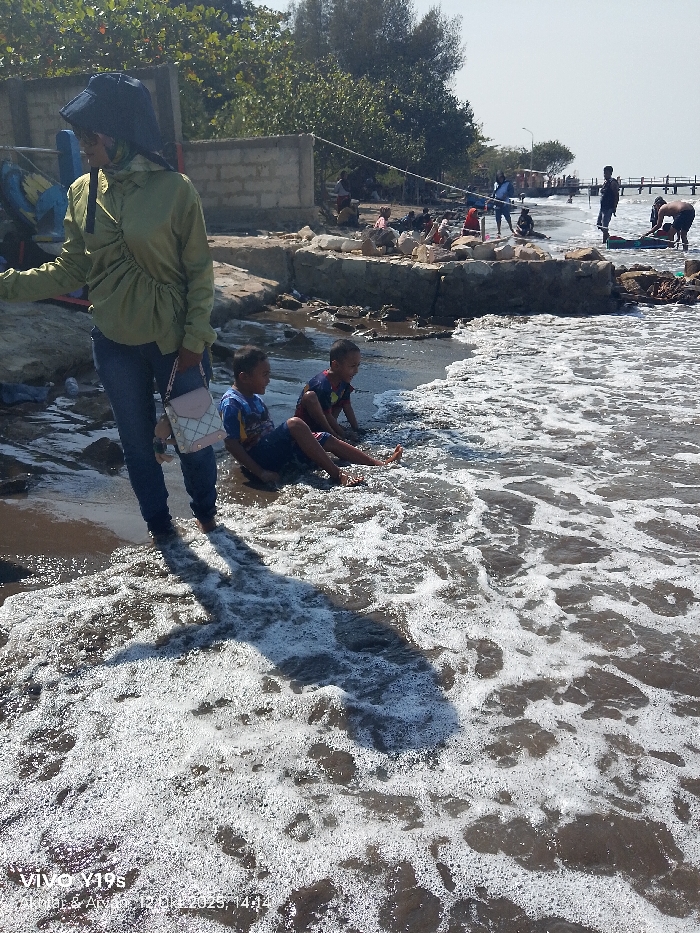 The image displays a coastal area in Limbangan, Indonesia, under clear, sunny conditions. In the foreground, foamy brown water covers a dark, sandy or muddy shoreline. An adult female stands on the left, dressed in a light olive-green long-sleeved top, dark blue jeans, and a dark bucket hat; a light pink and white patterned shoulder bag hangs from her left shoulder. Her feet are partially submerged, and a prominent dark shadow extends from her across the water. To her right, two young boys are seated in the shallow water; one wears a blue and yellow t-shirt, and the other a red, black, and yellow t-shirt, both observing the water. The midground features an uneven shoreline composed of concrete blocks, rubble, and dark earth, with scattered debris. Behind this barrier, green trees and a low concrete block wall are visible. Several indistinct figures are present on this elevated shoreline, engaged in various activities. The background shows an expanse of water leading to a wooden pier or jetty extending from the right, on which multiple people are standing. Additional individuals are visible along the distant shoreline, some near small boats.