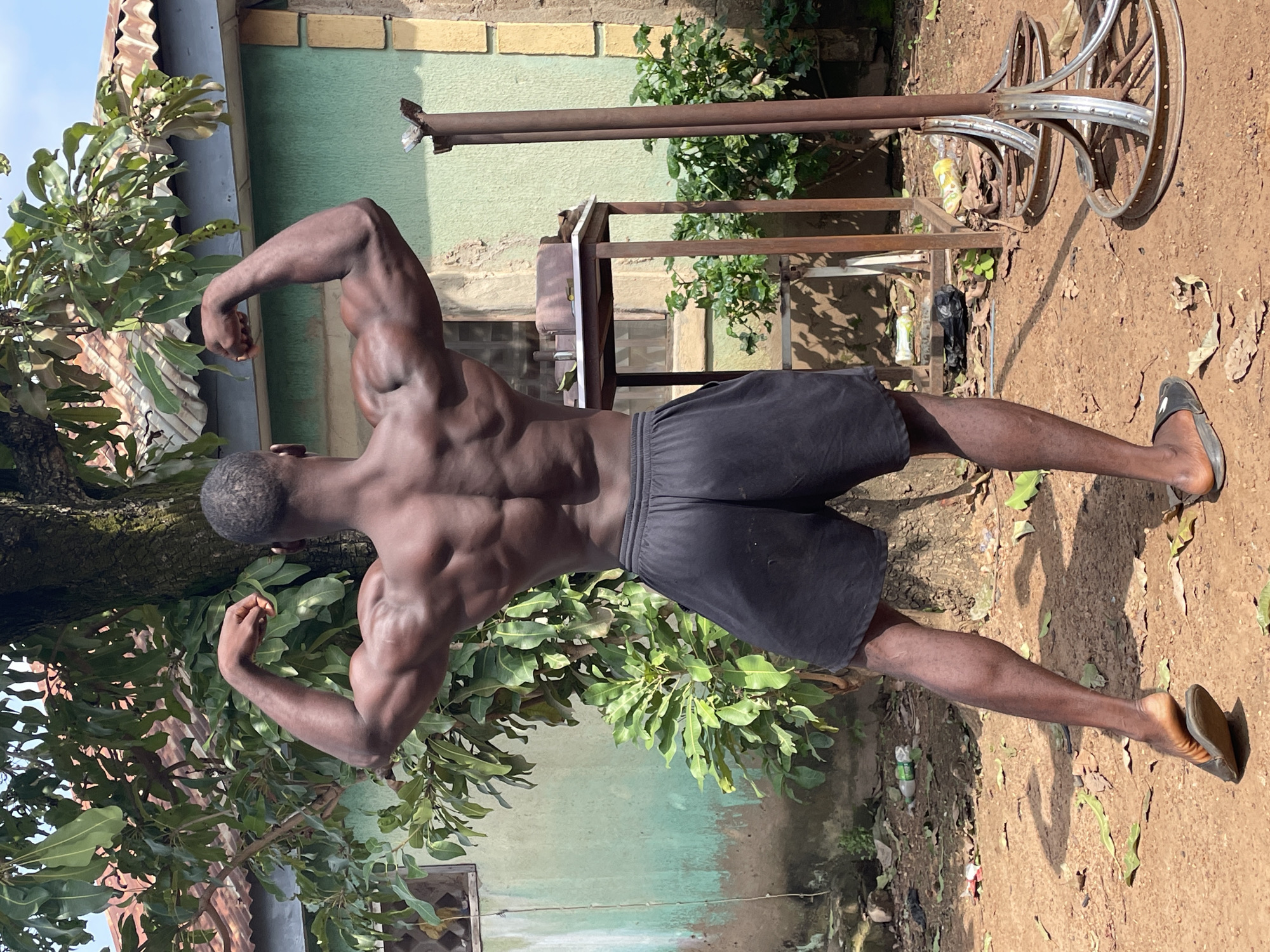 In Akwanga, Nigeria, a Black male subject stands outdoors, displaying his muscular back and arms. He is facing away from the camera, with his arms flexed upwards, showcasing his biceps and latissimus dorsi muscles. The subject is wearing black athletic shorts and flip-flops. The environment appears to be a somewhat unkempt outdoor area, possibly a backyard or a training space. Behind the subject, a tree with abundant green leaves is prominent. To the right, a light green wall of a building is visible, with yellow brick accents. In the background, there is a metal table with objects on top and some rustic metal structures resembling gym equipment. The ground is composed of dirt and scattered leaves. The lighting suggests it is daytime, with shadows cast on the ground.