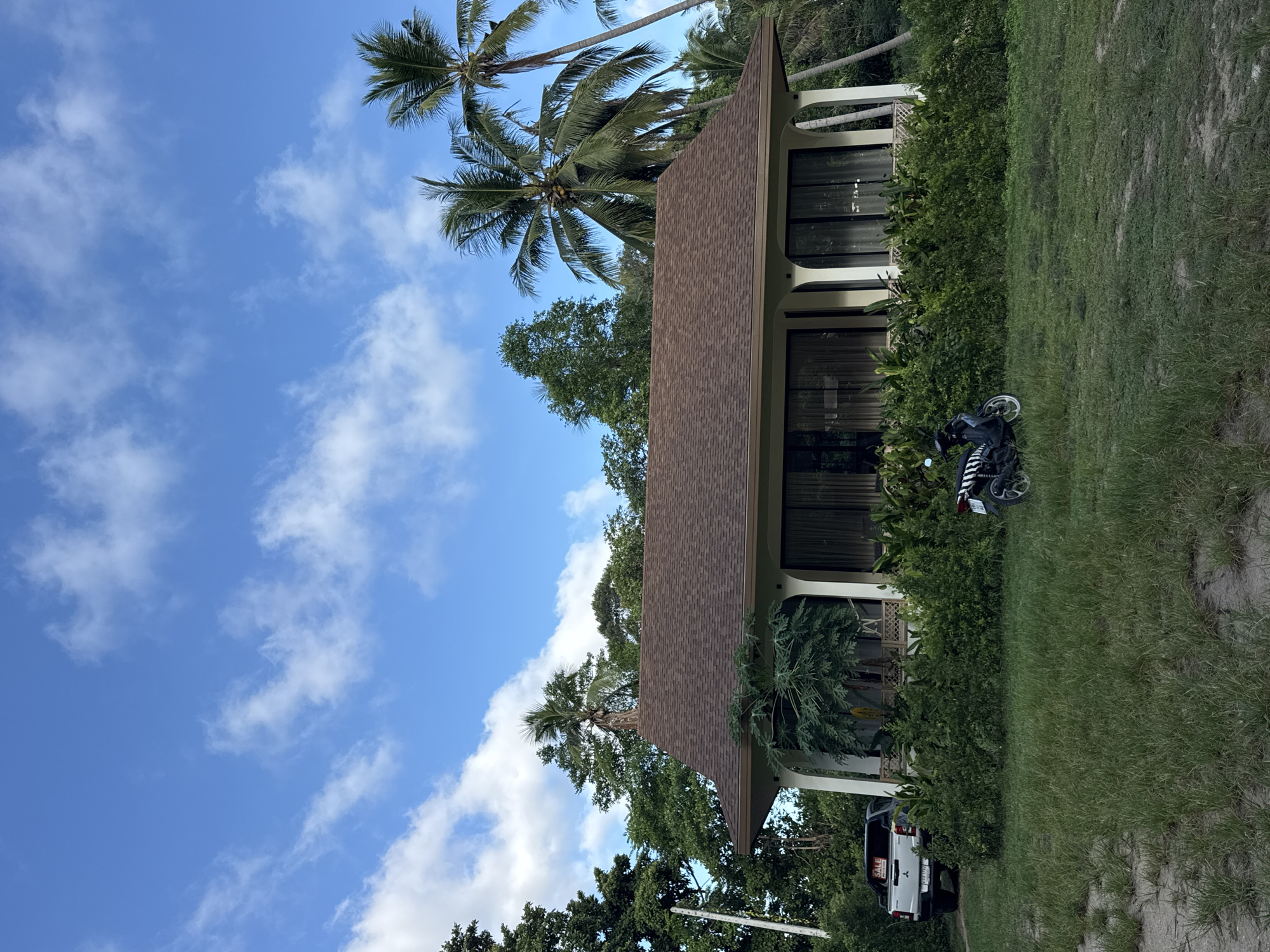 The image depicts a single-story house with a brown, tiled roof situated in a tropical environment. The house has large windows and white columns supporting the roof overhang. It is surrounded by lush green vegetation, including trees and bushes. Several palm trees are visible in the background, suggesting a coastal or island location. In front of the house, there is a patch of green grass. To the left of the house, a white pickup truck with a "SALE" sign on its rear window is parked on a dirt path. A black and white scooter is parked on the grass to the right of the house. The sky is blue with scattered white clouds. The scene appears to be in ตำบลเกาะพะงัน, Thailand, given the tropical vegetation and general ambiance. There are no people visible in the image.