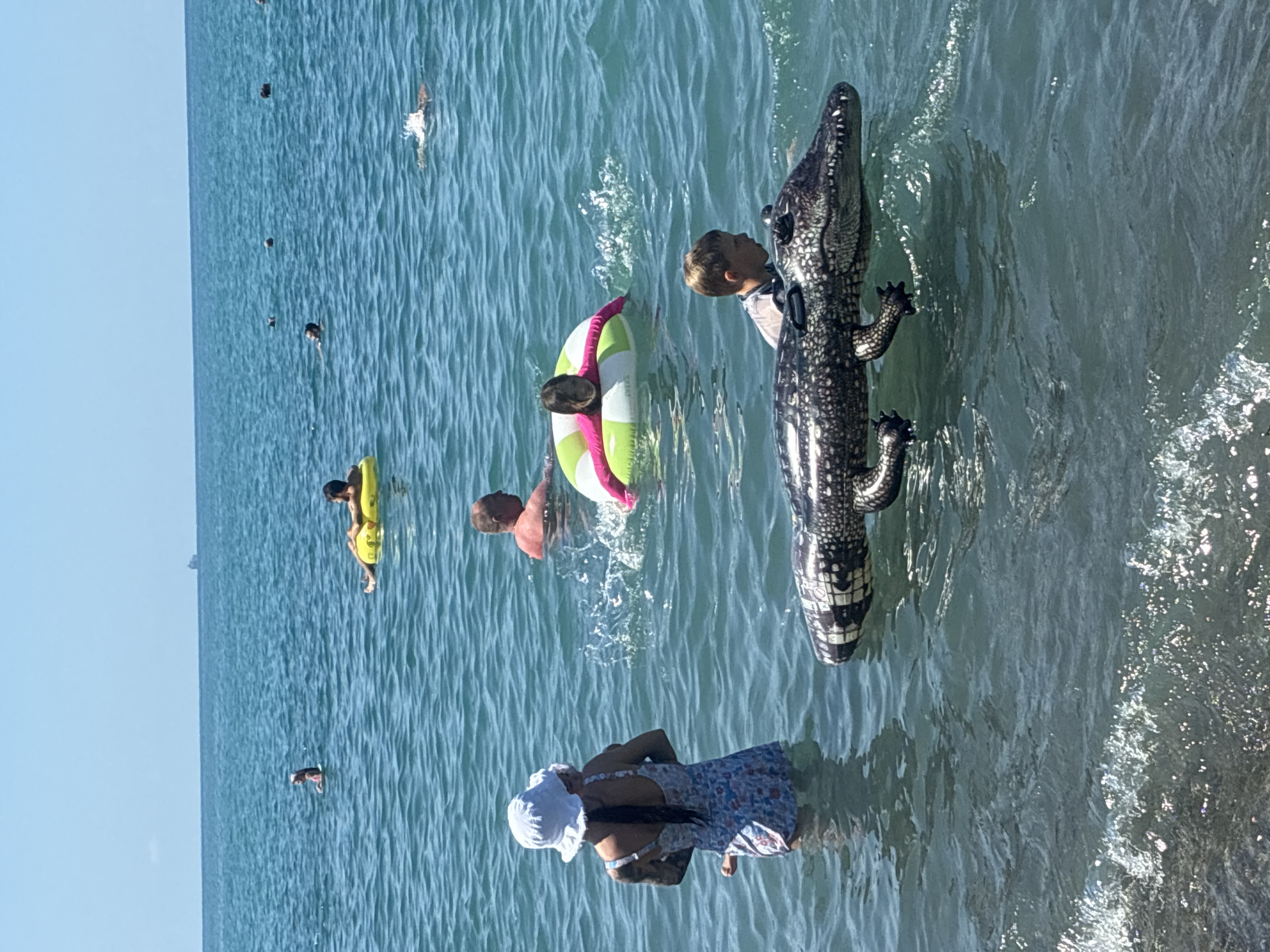 In Sochi, Russia, on a clear, sunny day, people enjoy the Black Sea. In the foreground, a large inflatable alligator floats in the water. A young boy is swimming near the alligator, and a woman wearing a white bucket hat and a floral swimsuit stands in the water, holding a baby. Further out, a girl in a pink and green inner tube is visible, as is a man floating on a yellow inflatable. Other swimmers are scattered across the blue water, and a distant cargo ship is visible on the horizon. The water is choppy, with small waves near the shore.