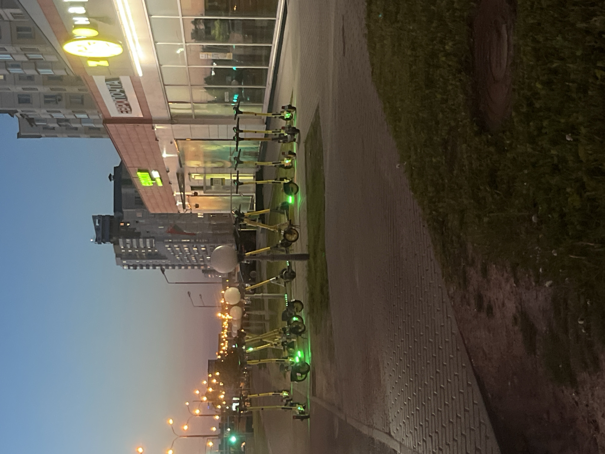 An evening urban street scene in Minsk, Belarus, is captured at dusk or early night, with artificial lights beginning to illuminate the environment. The image depicts a row of approximately ten yellow electric scooters (e-scooters) parked neatly along a paved sidewalk, adjacent to a multi-story building. Many of these scooters display distinctive glowing green lights, suggesting they are active or available for rental.
To the immediate right of the sidewalk is a narrow strip of well-maintained grass, where a dark, round manhole cover is partially visible. Further beyond, a road or open area extends into the distance, lined with numerous streetlights that emit a warm, golden glow. Some of these streetlights feature prominent white spherical lamps.
The building on the left, likely a commercial complex, features large glass storefronts that reflect parts of the street and sky. A prominent, horizontally oriented illuminated sign on this building displays the Cyrillic text "ЕВРООПТ" (YEVROOPT). Below this main sign, a white banner is visible with the Cyrillic text "ЕВРОПОЧТА" (YEVROPOCHTA).
The sky above is a clear, gradient blue, characteristic of twilight hours, gradually darkening towards the top of the frame. In the far background, other urban buildings are dimly visible against the fading light. No people are present, lending a quiet and calm atmosphere to the bustling city infrastructure.