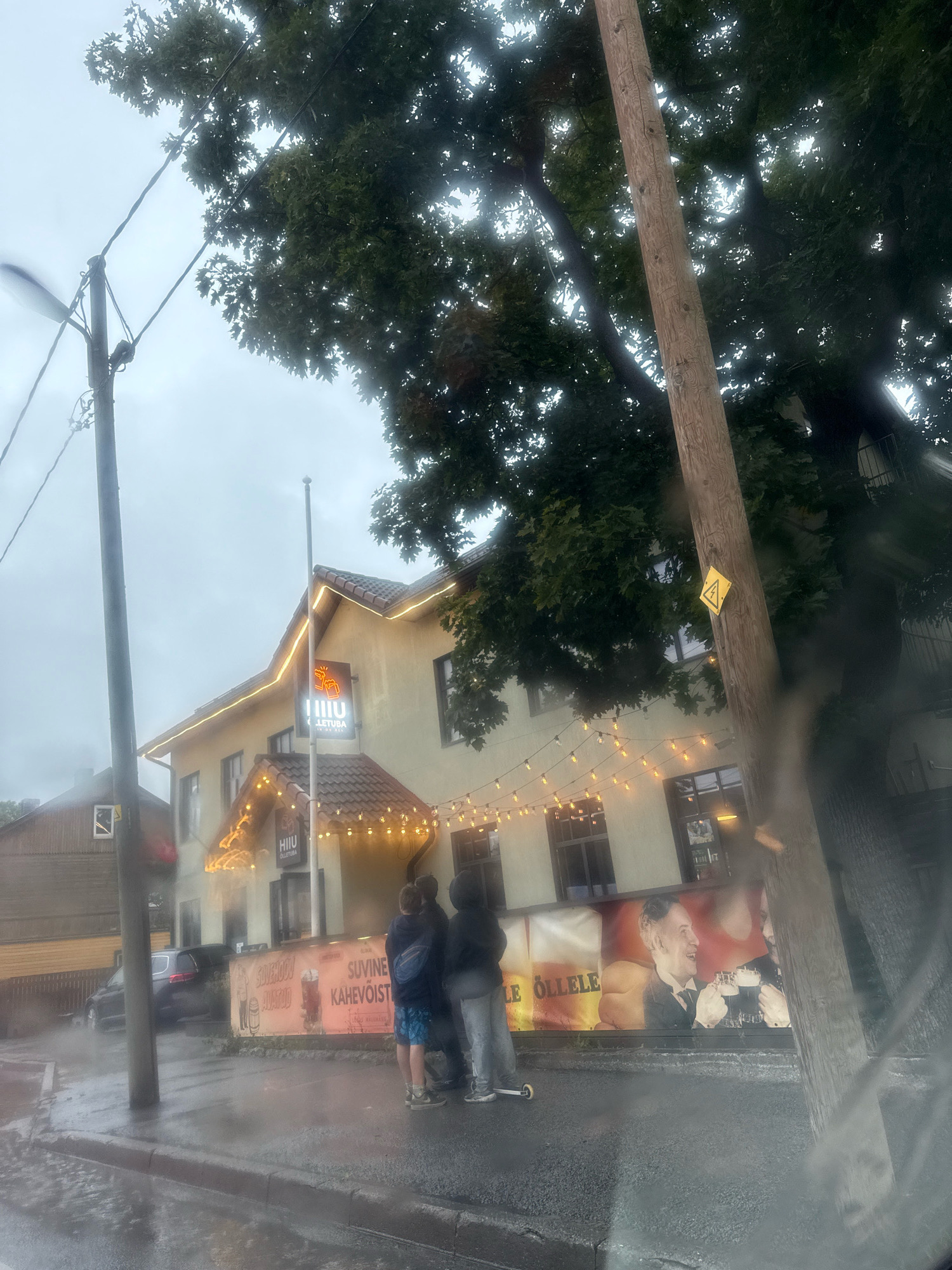 The image captures a rainy day scene on a street in Tallinn, Estonia, in front of a building with a sign that reads "HIIUMAA KÜLLATUBA". The building appears to be a restaurant or pub, decorated with string lights along its roofline and entrance. Several people are visible standing on the sidewalk, some appearing to be sheltering from the rain. A dark-colored car is parked on the street. A large banner on the building features images of two men toasting with beer and text that appears to be advertising beer or a beverage. A wooden utility pole with a yellow hazard sign depicting a lightning bolt is also prominently featured in the foreground, and a street lamp is visible to the left. The wet asphalt and reflections on the ground indicate recent or ongoing rainfall.