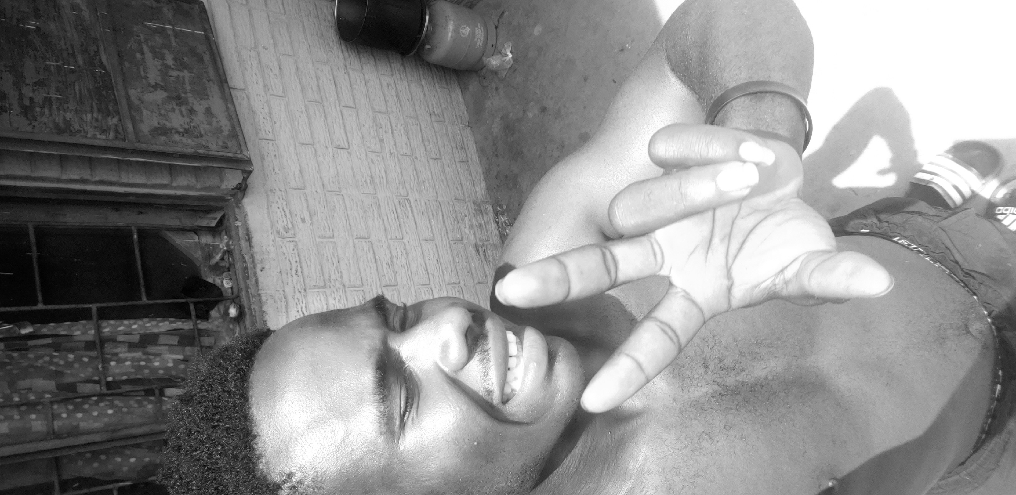 In Narayi, Nigeria, a close-up, grayscale, waist-up photograph captures a Black male in what appears to be an outdoor setting. The man is shirtless and has a short, textured haircut. He is looking to his right with his eyes closed and his mouth open, revealing his teeth in what seems to be a laugh or a grimace. His right arm is extended towards the camera, with his index and middle fingers held up in a peace sign. A dark, circular bracelet is visible on his left wrist. Behind him, a brick wall serves as a backdrop. To the far right, a portion of a gas cylinder with a black container on top is visible. The ground is paved and appears slightly wet. The scene suggests a candid moment, possibly in a residential area or an outdoor gathering space.