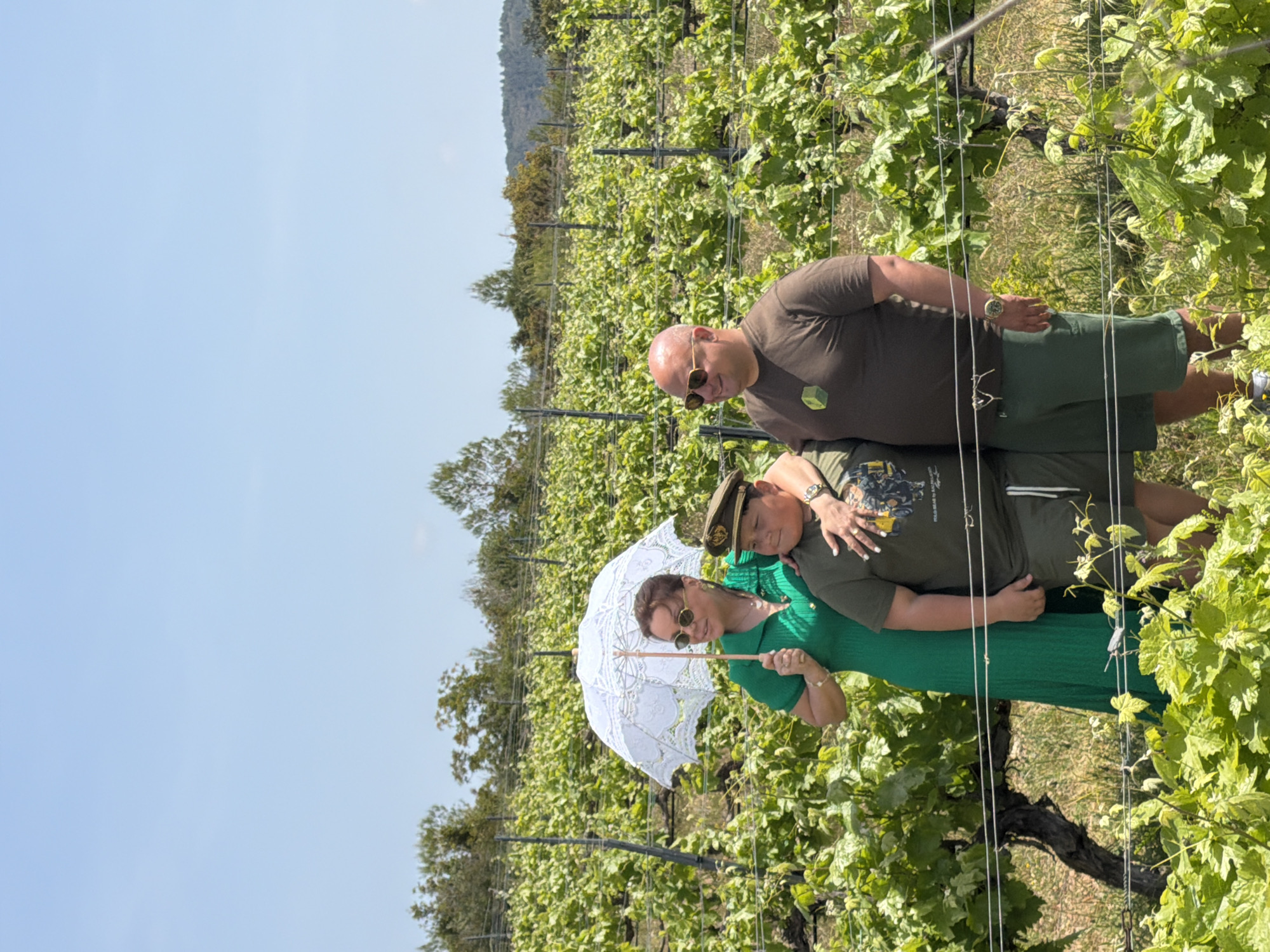 Three individuals, a man, a woman, and a child, are captured posing for a photograph in a sunny vineyard in Bonastre, Spain. The scene is bright and clear, with a vast blue sky overhead and rolling hills in the distant background.
In the foreground, rows of lush green grapevines with their leaves are visible. On the left, a bald man wearing sunglasses, a dark olive-green t-shirt, and shorts looks towards the camera with a slight smile. He wears a watch on his left wrist. To his right, a child, likely a boy, wearing a military-style cap and a similar dark olive-green t-shirt with an unidentifiable graphic print, leans affectionately against the woman. The woman, positioned on the right, wears a vibrant green dress and sunglasses. She holds a decorative white lace parasol over her shoulder, shielding herself from the sun. Her left arm is wrapped around the child, and a wedding ring is visible on her left hand. She also looks towards the camera with a subtle smile.
The trio appears to be enjoying an outing in the picturesque vineyard. The time of day is clearly daytime, given the bright sunlight. The atmosphere conveyed is relaxed and happy. No discernible text is clearly legible on any of the subjects' clothing or accessories.