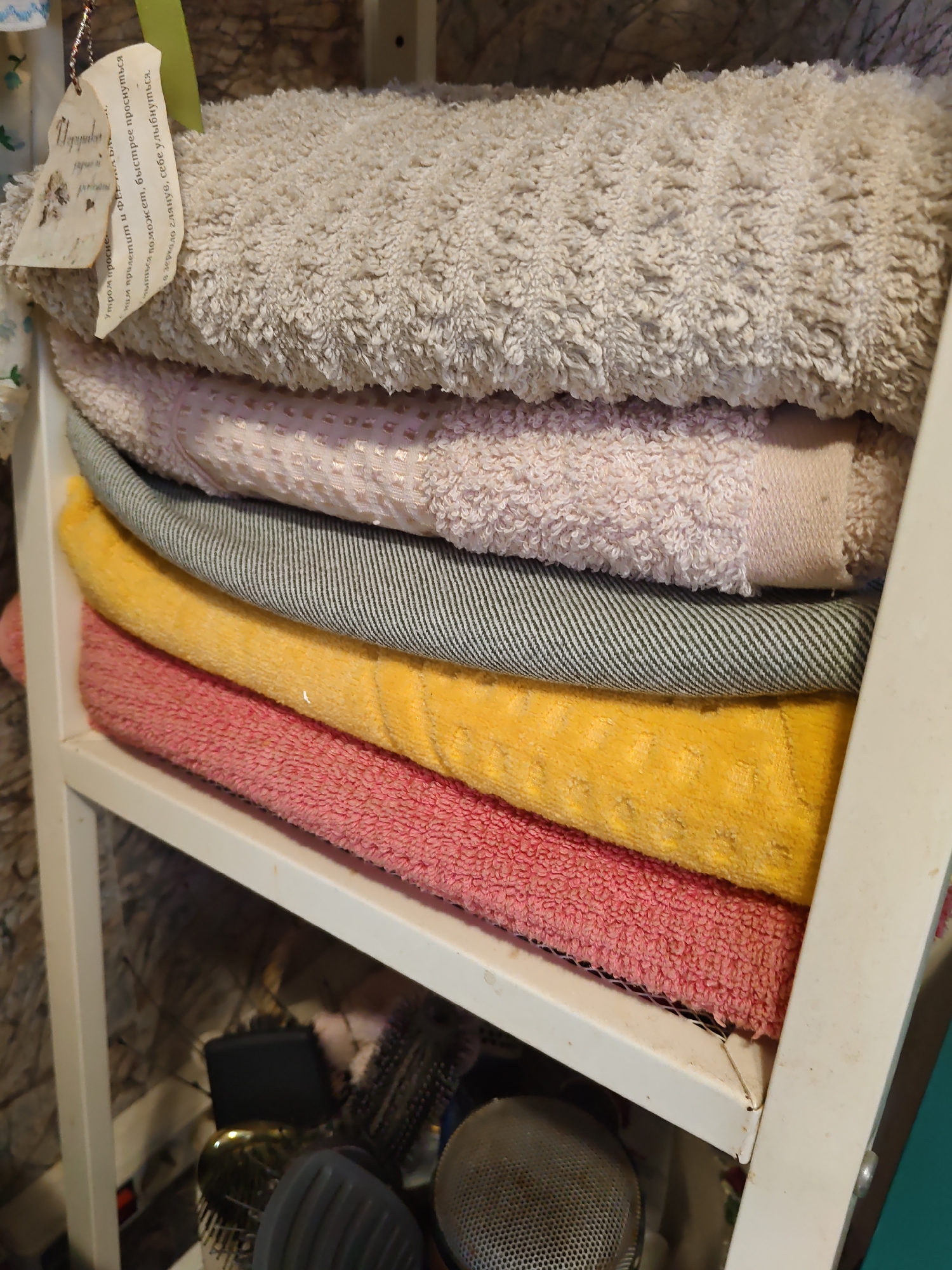 A stack of five folded towels is arranged on a white metal shelf. The towels, from top to bottom, are light beige, pale pink, grey with black stripes, bright yellow, and coral pink. The beige towel has a textured surface and a small, handmade tag attached to its corner. Below the towels, on the shelf, are various grooming tools, including brushes and what appears to be a black wallet or case. The background shows a mottled brown and grey surface, possibly a wall. The overall scene is a close-up interior shot, suggesting a bathroom or closet storage area. The image is undated and does not provide specific location information for Rostov-on-Don, Russia.