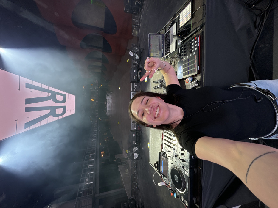 A female individual, appearing to be a DJ or performer, takes a selfie from a DJ booth inside the Mutabor club in Moscow, Russia. She stands behind a table covered in a black cloth, positioned center-frame, smiling broadly and making a peace sign gesture with her left hand while holding the camera with her right. She wears a black t-shirt, light-colored jeans, a black fanny pack, and in-ear monitors. A distinct black, segmented line tattoo is visible on her right forearm.
The DJ booth features a professional setup, including a Pioneer CDJ-3000 multi-player and a Pioneer DJM mixer on the left, a MacBook Pro laptop, an Akai MPC One, another grid-based MIDI controller (possibly an Ableton Push), and multiple microphones on the right.
The environment is a large, multi-level music venue with an industrial aesthetic. The background shows a vast, mostly empty dance floor and tiered viewing platforms on both sides, suggesting a capacity for a substantial audience. Minimal scattered individuals are visible on the lower level. Overhead, a large, illuminated trapezoidal screen displays the mirrored letters "RR." Haze permeates the upper air, illuminated by spotlights from the ceiling. Distinctive red, curved architectural elements are visible along the right wall of the venue.