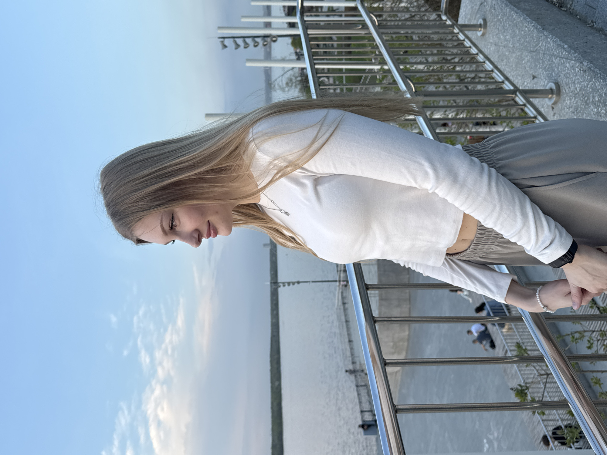 A young woman with long blonde hair stands on a metal railing overlooking a body of water. She is wearing a white long-sleeved crop top and grey pants. Behind her is a concrete structure with metal railings. In the background is a body of water with structures on the opposite shore. Based on the location, it is likely in Perm, Russia, along the Kama River embankment.