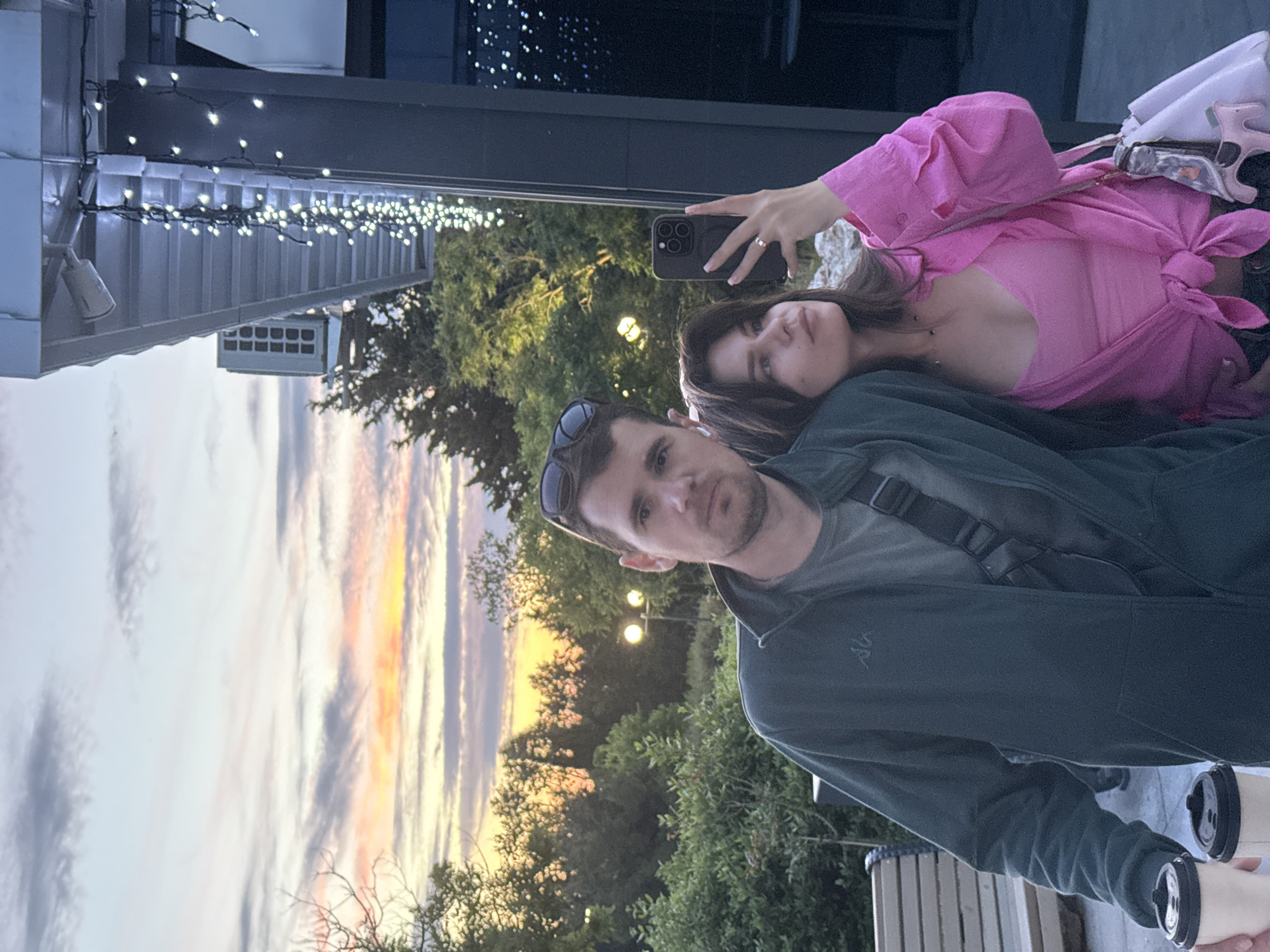 A young couple takes a selfie outdoors at dusk. The man, on the left, wears a dark green zip-up jacket over a grey t-shirt and has sunglasses perched on his head. He holds two disposable coffee cups in his left hand. The woman, on the right, wears a pink crop top and a pink button-up shirt tied at her waist. She holds a black smartphone in her right hand, capturing the selfie. A light pink handbag is slung over her left shoulder. The background features lush green trees and a colorful sunset sky with streaks of orange, pink, and grey clouds. To the right, a dark building with string lights and an outdoor air conditioning unit is visible. The overall setting appears to be a cafe or restaurant patio.