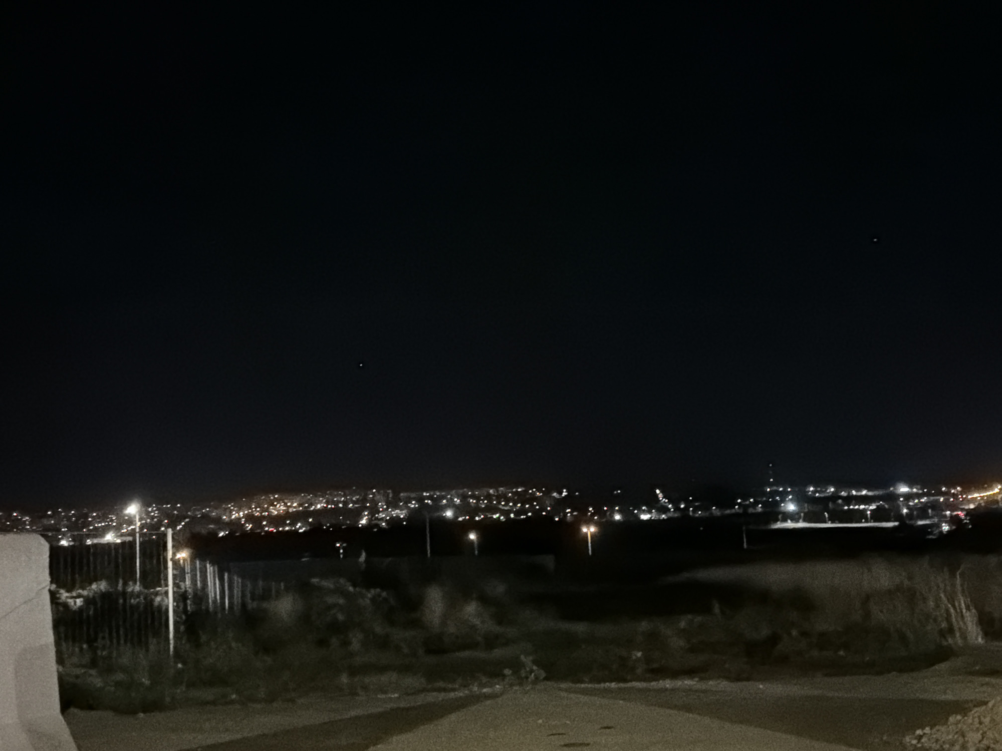 The image depicts a nighttime view of the city of Nitra, Slovakia. In the foreground, a dark, unpaved path leads towards a densely populated urban area illuminated by numerous lights. The city lights stretch across the horizon, indicating a significant settlement. The sky above is dark, with two faint celestial bodies visible. A fence line, possibly a border or barrier, is discernible in the middle ground, separating the foreground from the city. The overall scene is one of urban sprawl at night, viewed from a slightly elevated position.