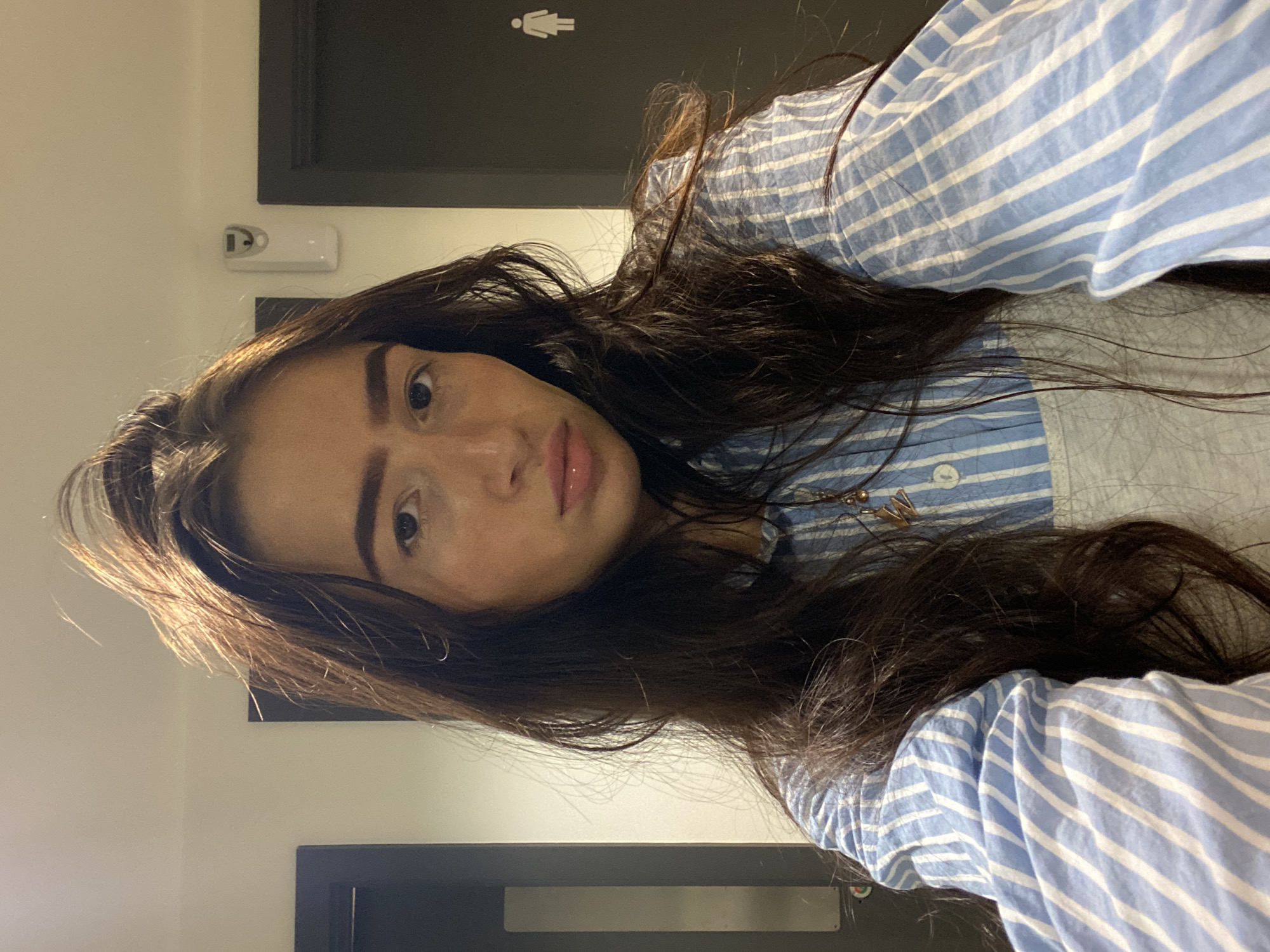 A young woman with long brown hair and light skin is posing for a selfie. She is wearing a blue and white striped shirt. Behind her is a dark-colored door with a white female restroom symbol and a white rectangular object mounted on the wall to the left. A portion of a white wall is also visible. There is not enough information to determine the specific location in the city of London.