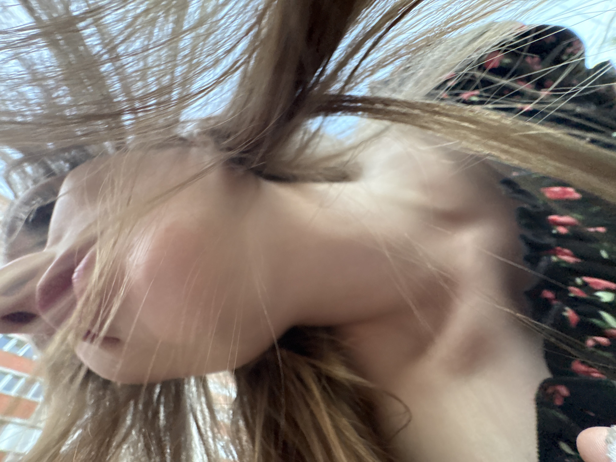A dynamic, low-angle close-up photograph captures a person, likely a young female, from a perspective looking upwards. The subject's face is partially obscured by a cascade of light brown or blonde hair, which appears to be in motion, possibly wind-blown or tossed.
The person's head is tilted, with only the chin, jawline, and a portion of the mouth with pinkish lips visible on the left side of the frame. Her gaze seems to be directed downwards or away from the camera. She is wearing a dark garment, possibly a dress or top, adorned with a repeating pattern of small red and pink floral motifs, visible on the right side of the image.
The background features a bright, light blue sky, indicating an outdoor setting during daytime. Faintly visible behind the person's head are structural elements of a building with multiple rectangular windows, suggesting an urban or residential environment, consistent with the Izhevsk, Russia location context. The lighting is bright and natural. No visible text is present in the image. The overall impression is candid and energetic, highlighted by the movement of the hair.