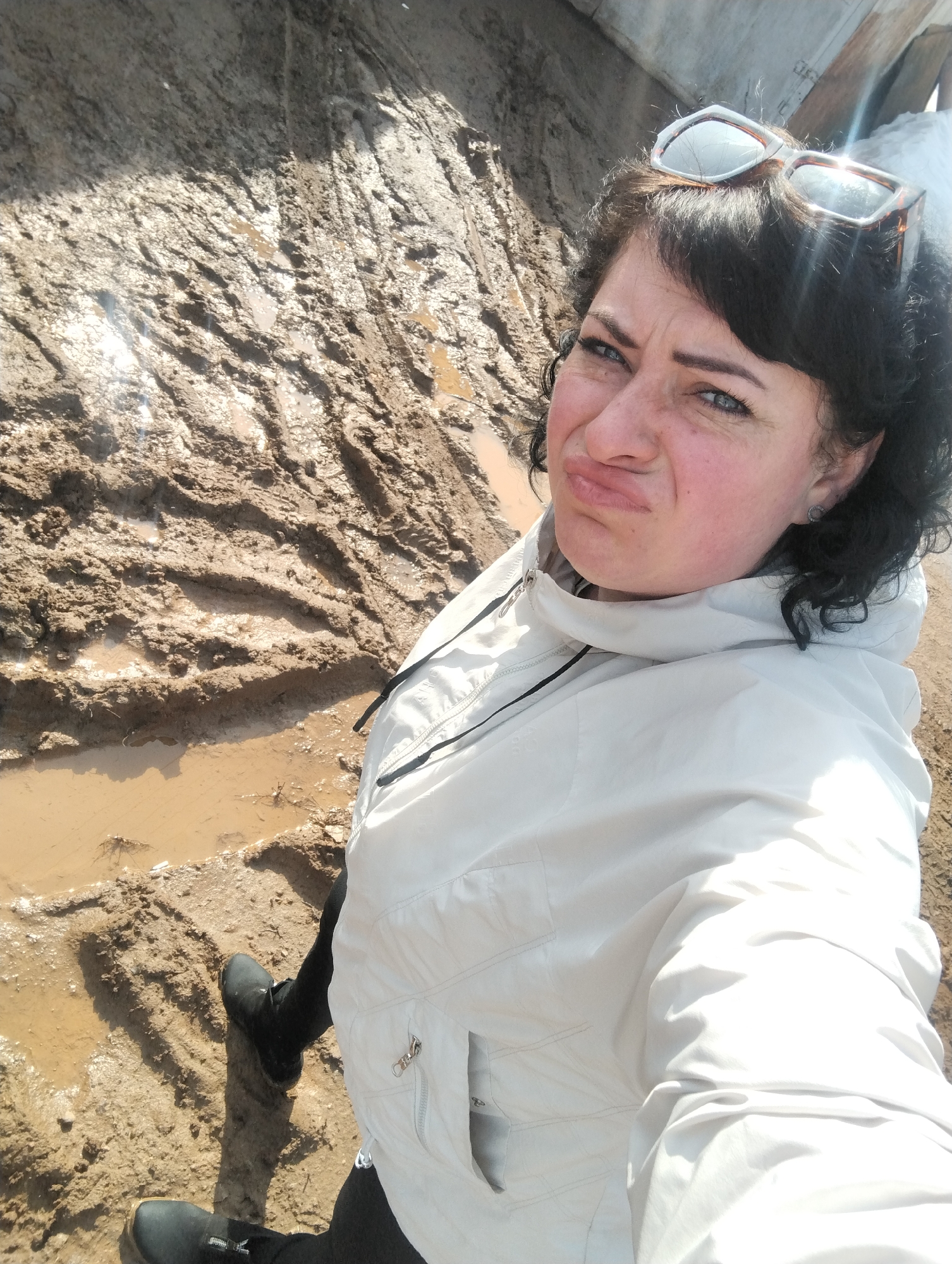 A woman is taking a selfie in a muddy environment. The ground is covered in thick mud with visible tire tracks and puddles. The woman is wearing a light-colored jacket, black pants, and black boots. She has sunglasses on her head and is making a pouting face. The location is Grishanki, Russia.