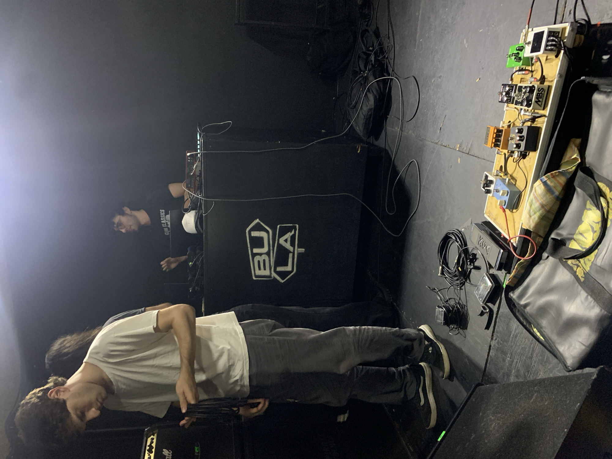 A dim and somewhat tilted medium shot shows two men and music equipment on a stage or platform in what appears to be a dark venue. The man in the foreground is facing towards the right, holding an instrument. He is wearing a white t-shirt, dark jeans, and dark shoes. Behind him is a taller black amplifier with white lettering saying "BULA". In the background, another man, possibly also a musician, is positioned near a DJ setup, partially obscured by the amplifier. A pedalboard with multiple effects pedals sits on the floor, alongside cables, adapters, and a large black bag with some yellow accents. The environment is dark, and the lighting appears to be stage lighting. The image includes enough elements to suggest this might be a music venue in Buenos Aires, Argentina.