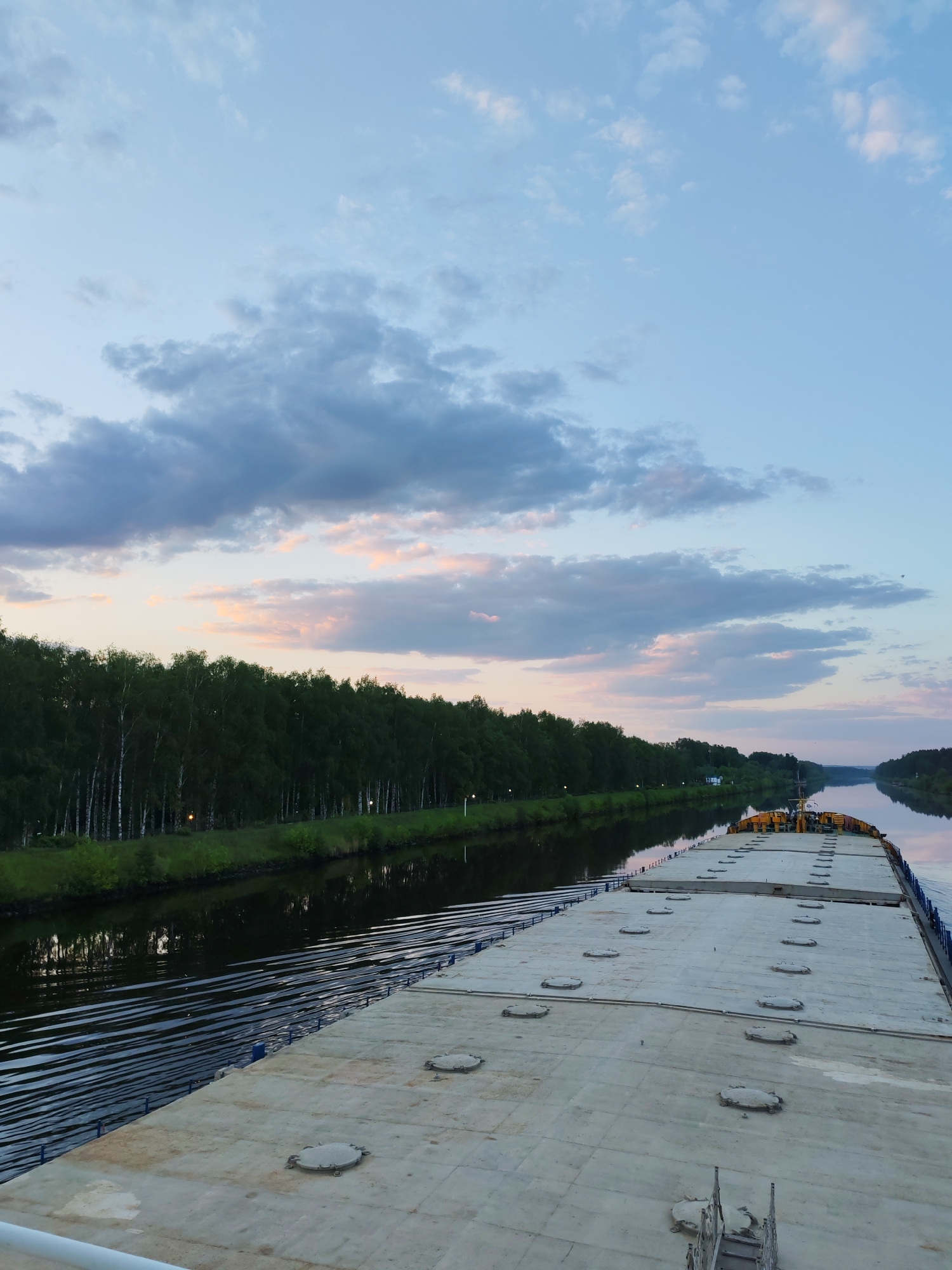 A broad, flat-decked vessel travels along a straight, narrow waterway in Татищево, Russia. The vessel's light gray deck is punctuated by circular access hatches. A line of trees lines the left bank of the waterway, their reflection mirrored in the dark water. A light-colored sky with streaks of pink and gray clouds dominates the upper half of the frame. The time is evening.