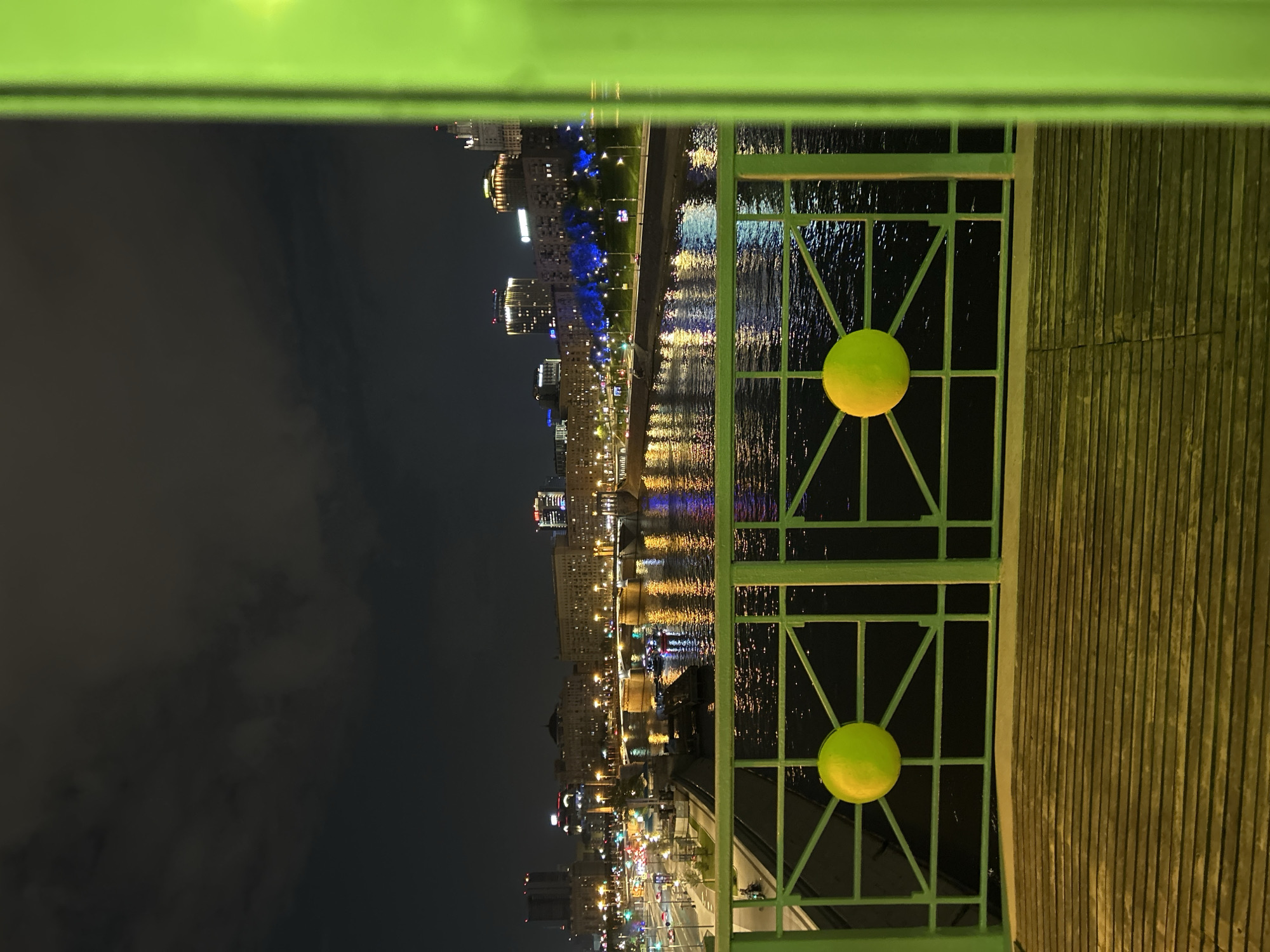 The image depicts a nighttime cityscape, potentially of Unknown, Unknown. A green metal railing with two large, yellow, spherical ornaments is prominently featured. The background reveals a river reflecting the lights of buildings and infrastructure along the embankment. The sky is dark, with hints of clouds. The scene suggests an urban environment viewed from a bridge or elevated walkway.