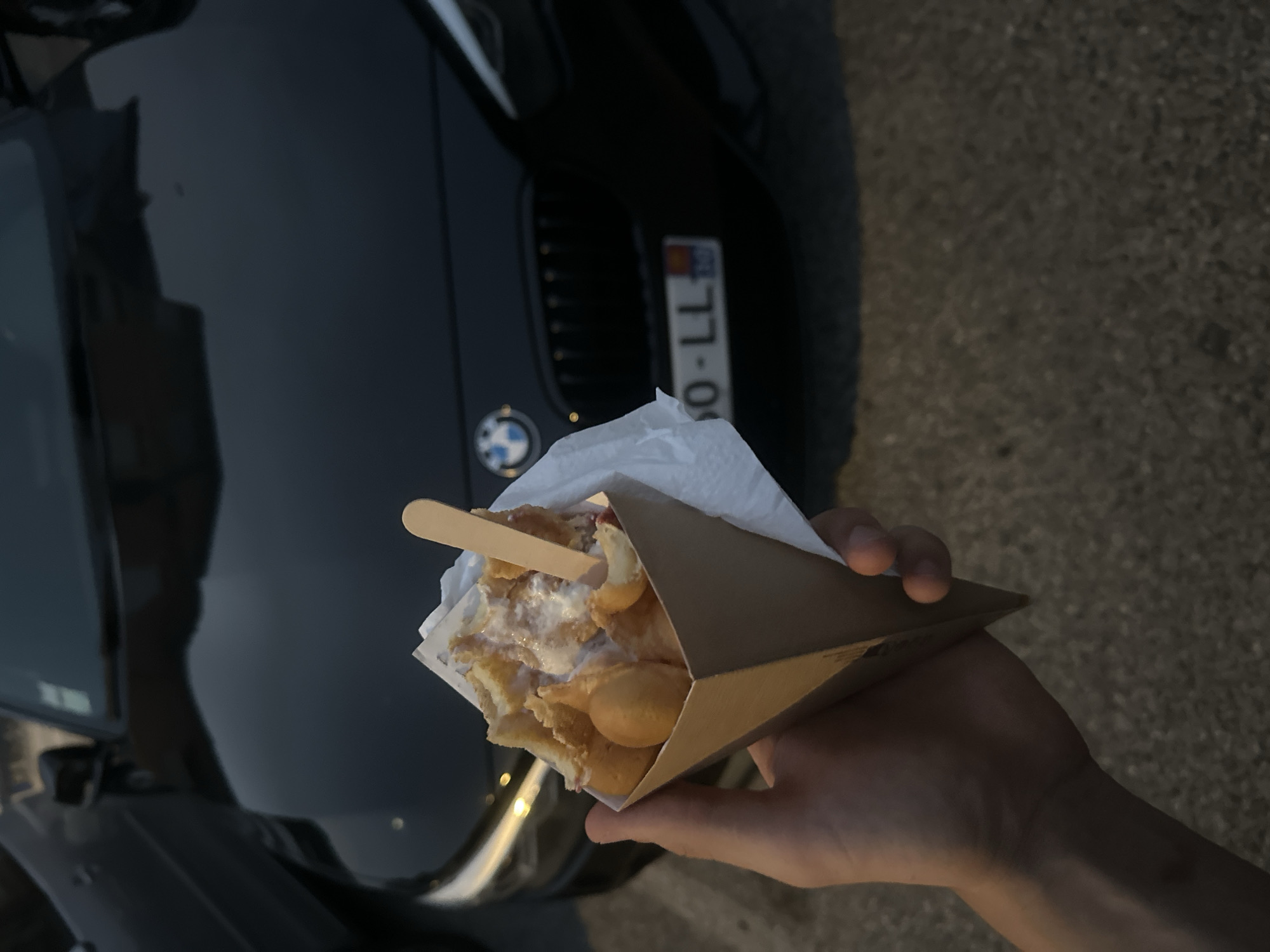 A first-person perspective shot captures a person holding a waffle cone filled with a dessert in front of a black BMW. The waffle cone is made of brown paper and is filled with a light-colored dessert, possibly ice cream or custard, with some fruit visible. A wooden popsicle stick is inserted into the dessert. The BMW is parked behind the cone, with its grille, headlights, and license plate visible. The license plate appears to have a European format, with "60-LL" and some other characters. The ground in the foreground is paved and textured. The lighting suggests it is either early morning or late evening, with warm, soft light.