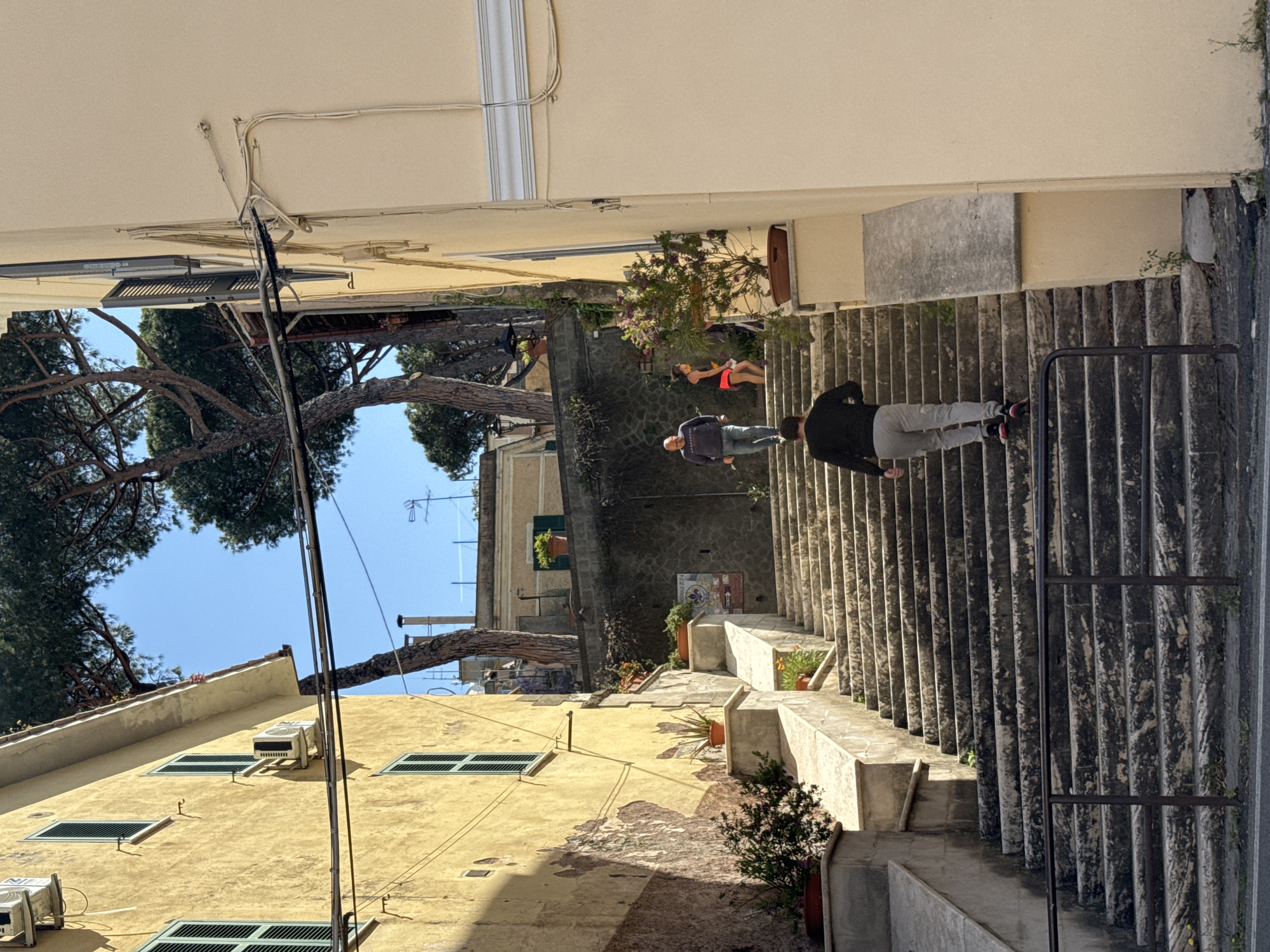 This image captures a vibrant, sunlit stepped alleyway in Castel Gandolfo, Italy, descending between old buildings. The scene is dominated by concrete stairs and landings, bordered by textured stone walls and railings.
Two individuals are visible walking down the main staircase. In the immediate foreground, a person with their back to the viewer wears a black top, grey sweatpants, and sneakers with red accents, holding a phone. Further down, another man in a dark grey jacket and light jeans also walks down the steps. On a flatter section between staircases, a person in an orange top and red shorts is seen resting or standing.
The buildings lining the alley are primarily painted in a warm, muted yellow, showing signs of age with some crumbling plaster. They feature several windows, some fitted with green-slatted shutters, and multiple white air conditioning units. Exposed electrical conduits and wires are visible running along the building facades and under overhangs.
Large, mature pine trees, likely umbrella pines, frame the bright blue sky in the upper left, their dense foliage providing shade. Numerous potted plants with greenery and some flowers are arranged along the stair landings and ledges, adding splashes of color. A dark metal railing runs along the right side of the main staircase. On one stone wall, a small, colorful mosaic artwork is visible, which appears to have text, possibly including "PALAZZO" and "2014", though it's not perfectly clear.
The strong, distinct shadows cast by the buildings and trees indicate a clear, sunny mid-day. The overall atmosphere is one of a charming, historic Mediterranean town.