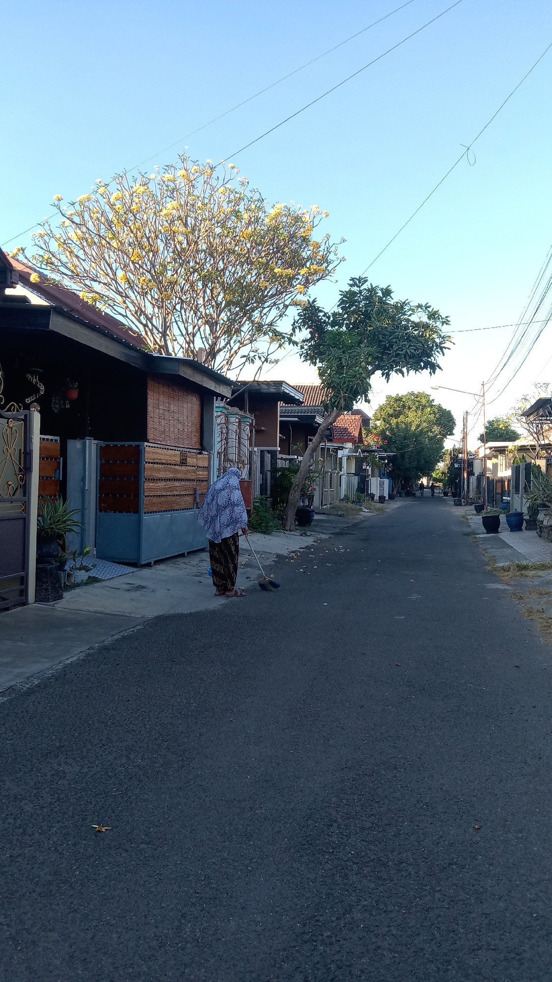 A person wearing a headscarf and patterned skirt sweeps the asphalt road in front of a row of houses in Kediri City, Indonesia. The sun is shining, casting shadows on the road. A large tree with yellow flowers is in the foreground, and other trees and houses line the street. Power lines are visible in the sky. The scene is peaceful and ordinary.