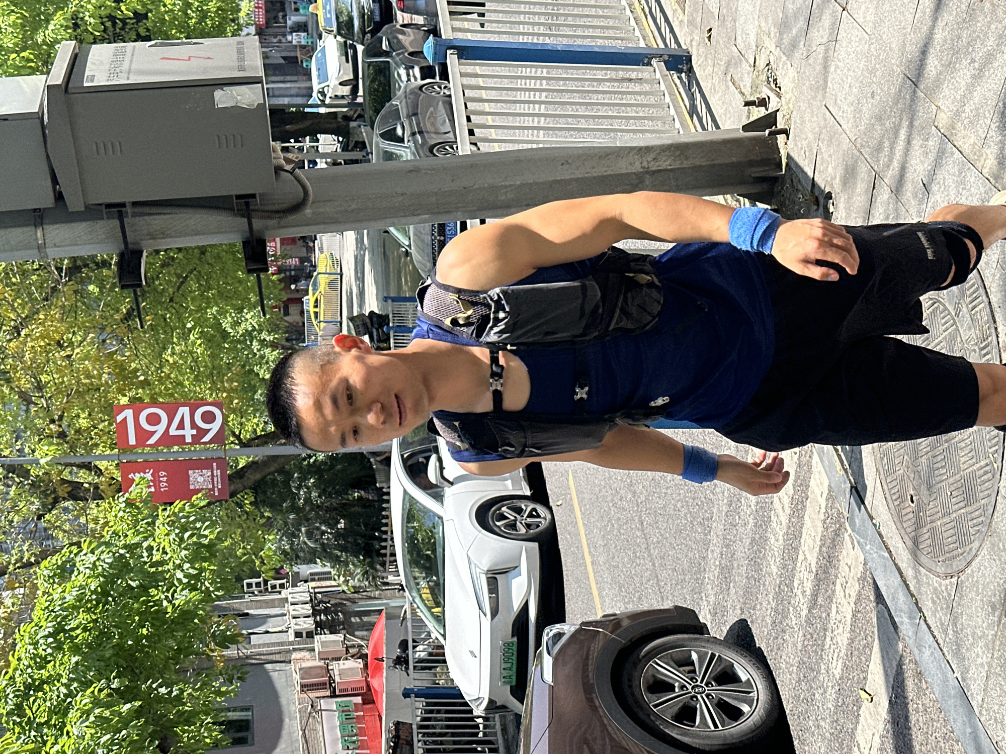 A medium shot presents a man standing on a sidewalk, clad in a blue shirt, black shorts, and blue wristbands. He wears a black chest pack. Adjacent to him is a street with cars, including a white car with license plate A.AJ9098. Street signage with the number "1949" is visible. The location appears to be in Shapingba District, China, indicated by the street signage and license plate. The sidewalk is tiled with a manhole cover.