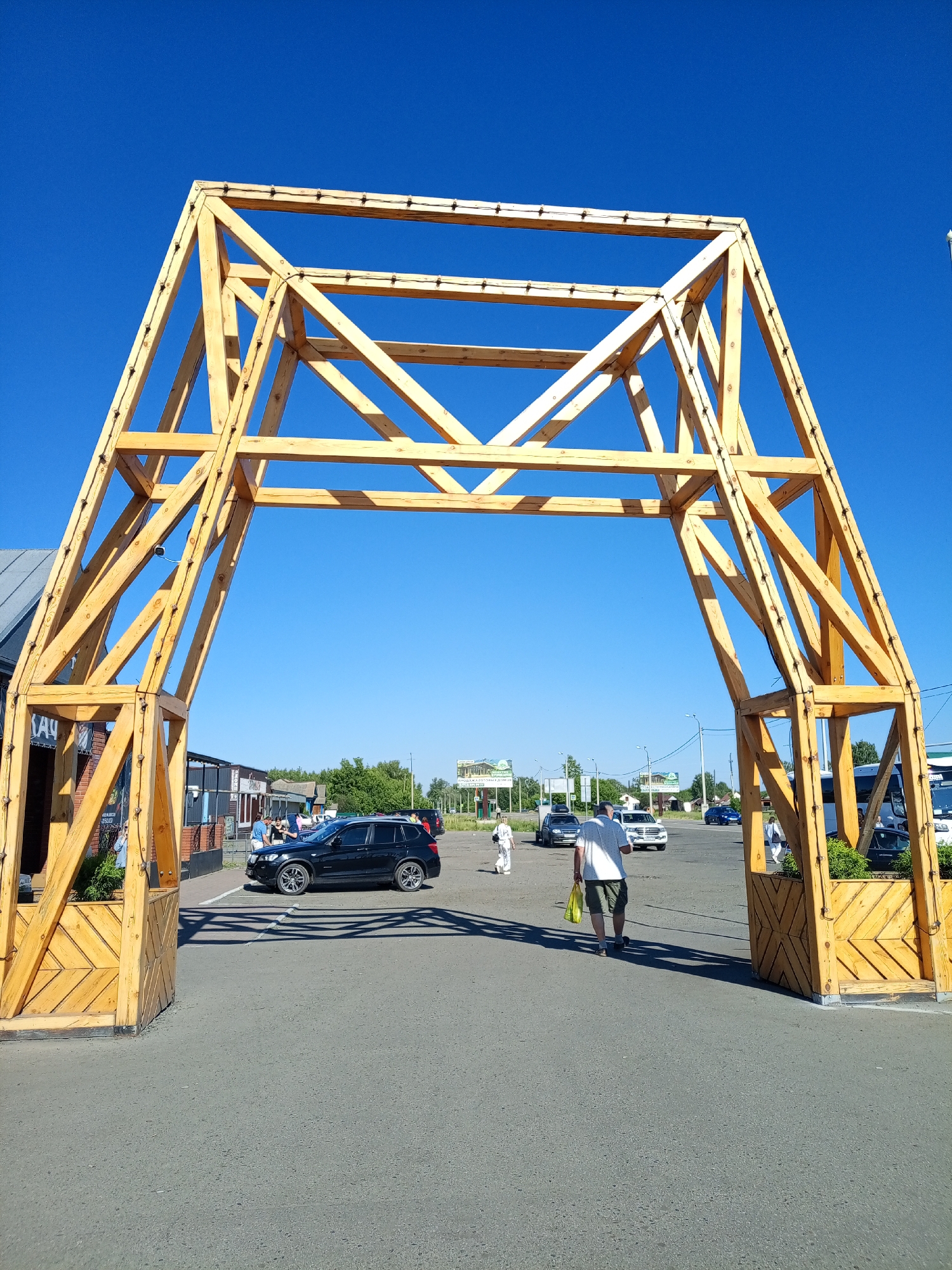 In Srostki, Russia, a large, wooden archway stands on a paved surface under a clear blue sky. The archway is constructed from light-colored wood beams forming an intricate, lattice-like structure. Small lights are strung along the upper beams of the arch. On the left side of the arch, there is a building with a sign that has Cyrillic writing. Several cars are parked and driving in the area behind the arch. A black SUV is parked on the left, and a white van or bus is visible further back on the right. People are scattered around the scene, some walking, and others standing near the buildings. One man in a white shirt and shorts is walking away from the camera carrying a yellow bag. The ground is asphalt, and shadows from the archway are cast across it. Greenery is visible in planters at the base of the archway and in the distance. The overall impression is an outdoor public space, possibly a marketplace or entrance to a facility.